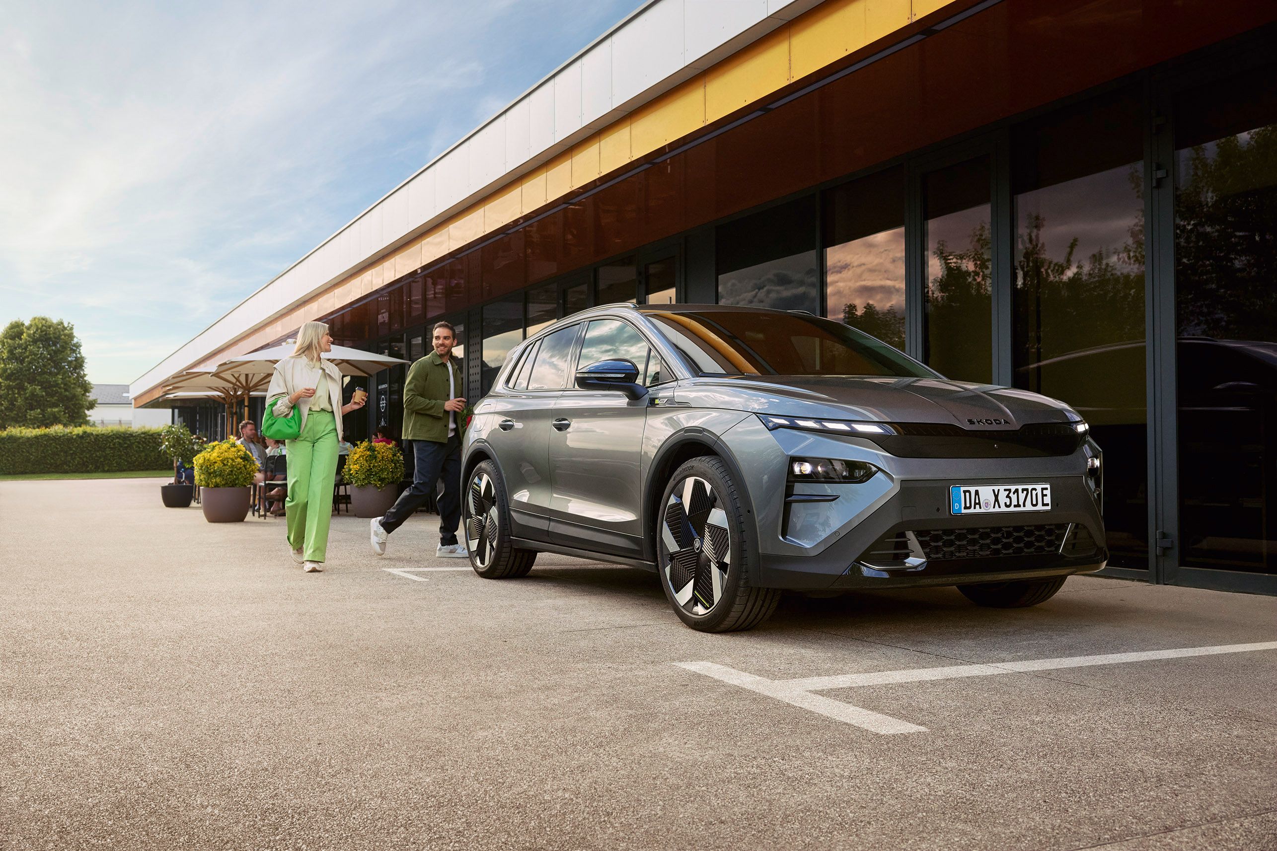 Na parkirišču ob sodobni stavbi je parkiran sodoben SUV z elegantno obliko. V ozadju sta dve osebi, ki se sprehajata po terasi z mizami in sončniki.