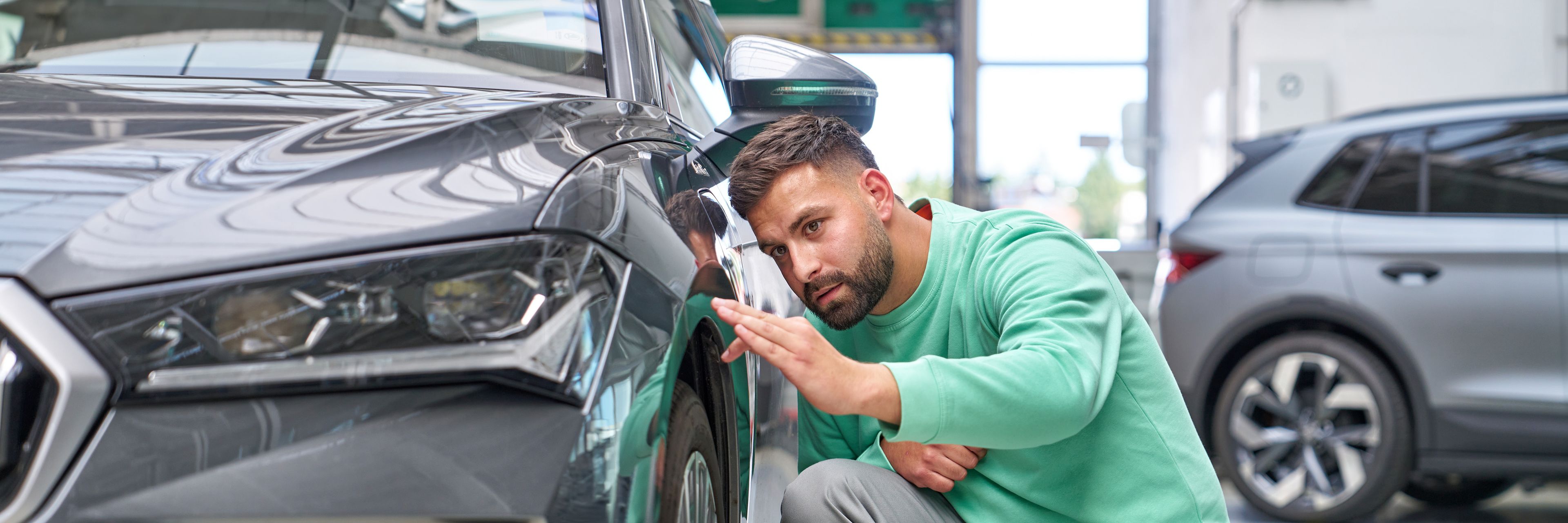 Oseba, oblečena v zeleno majico in sive hlače, pregleduje sprednje kolo črnega avtomobila. Avto je parkiran v svetli garaži, v ozadju je še en avtomobil.