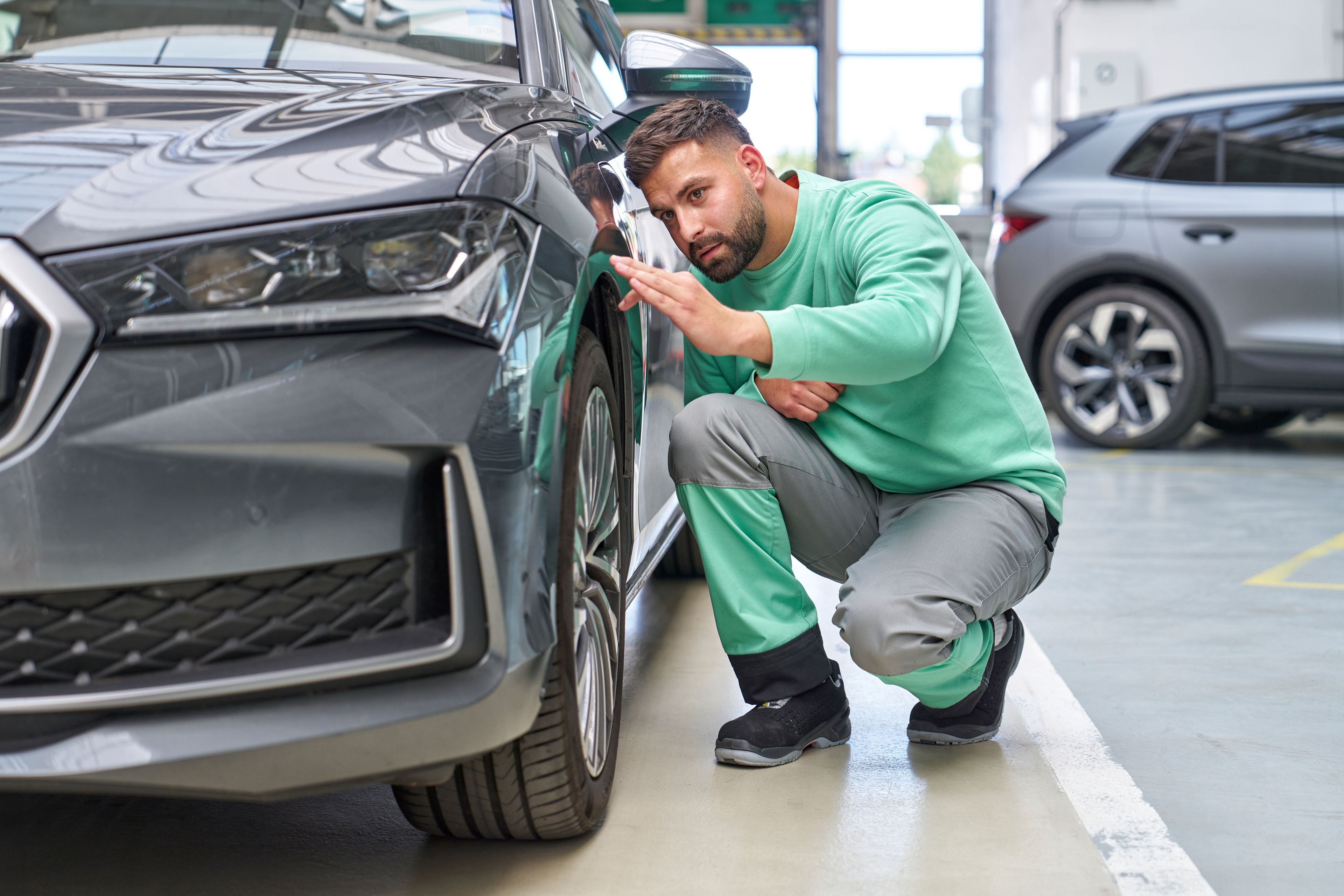 Oseba, oblečena v zeleno majico in sive hlače, pregleduje sprednje kolo črnega avtomobila. Avto je parkiran v svetli garaži, v ozadju je še en avtomobil.