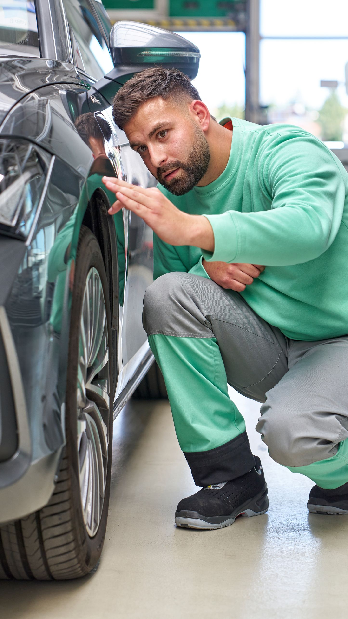 Oseba, oblečena v zeleno majico in sive hlače, pregleduje sprednje kolo črnega avtomobila. Avto je parkiran v svetli garaži, v ozadju je še en avtomobil.