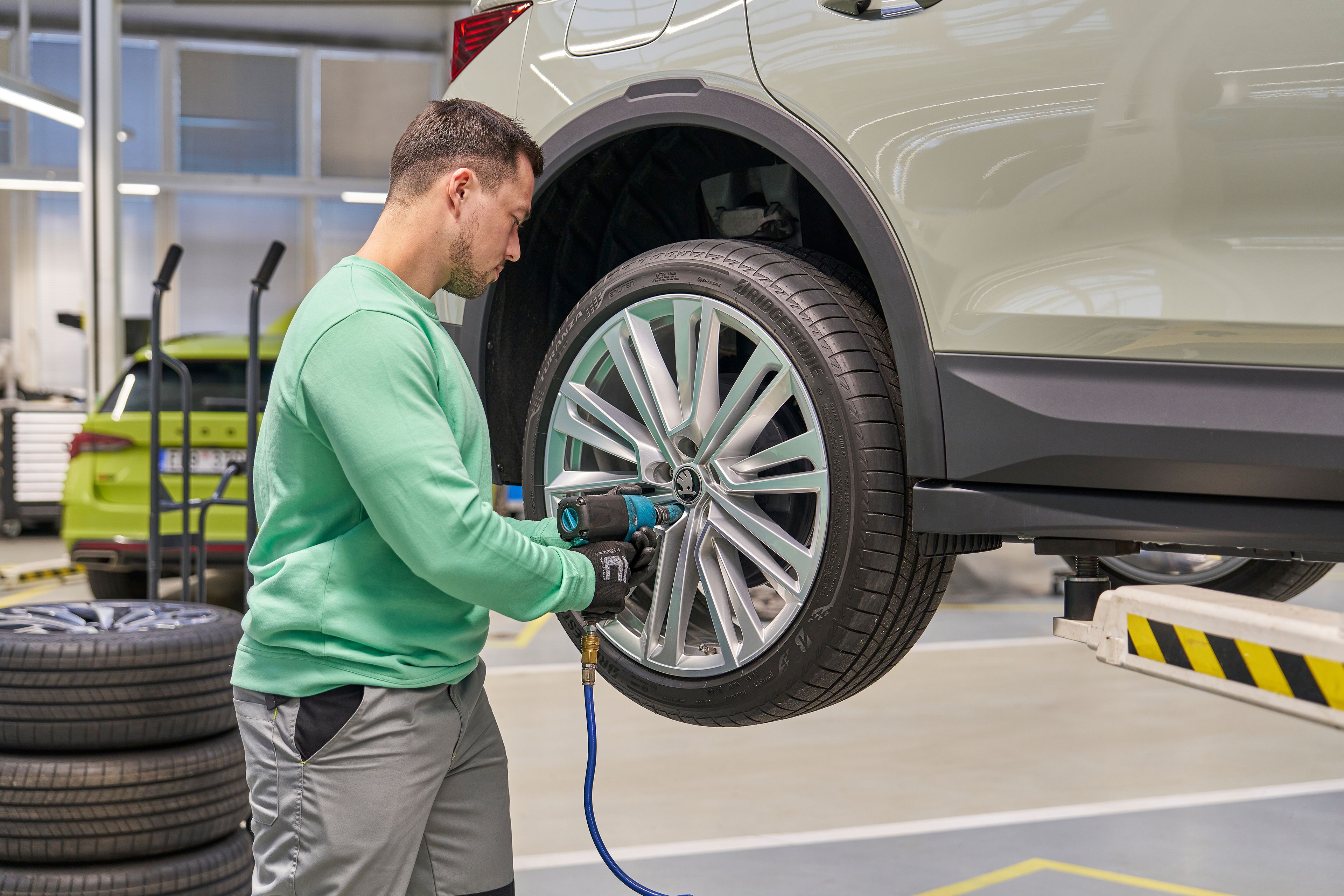 Oseba v delovni uniformi zamenjuje pnevmatiko na avtomobilu, dvignjenem na dvigalki. V ozadju so vidne dodatne pnevmatike in drugo vozilo v avtomehanični delavnici.