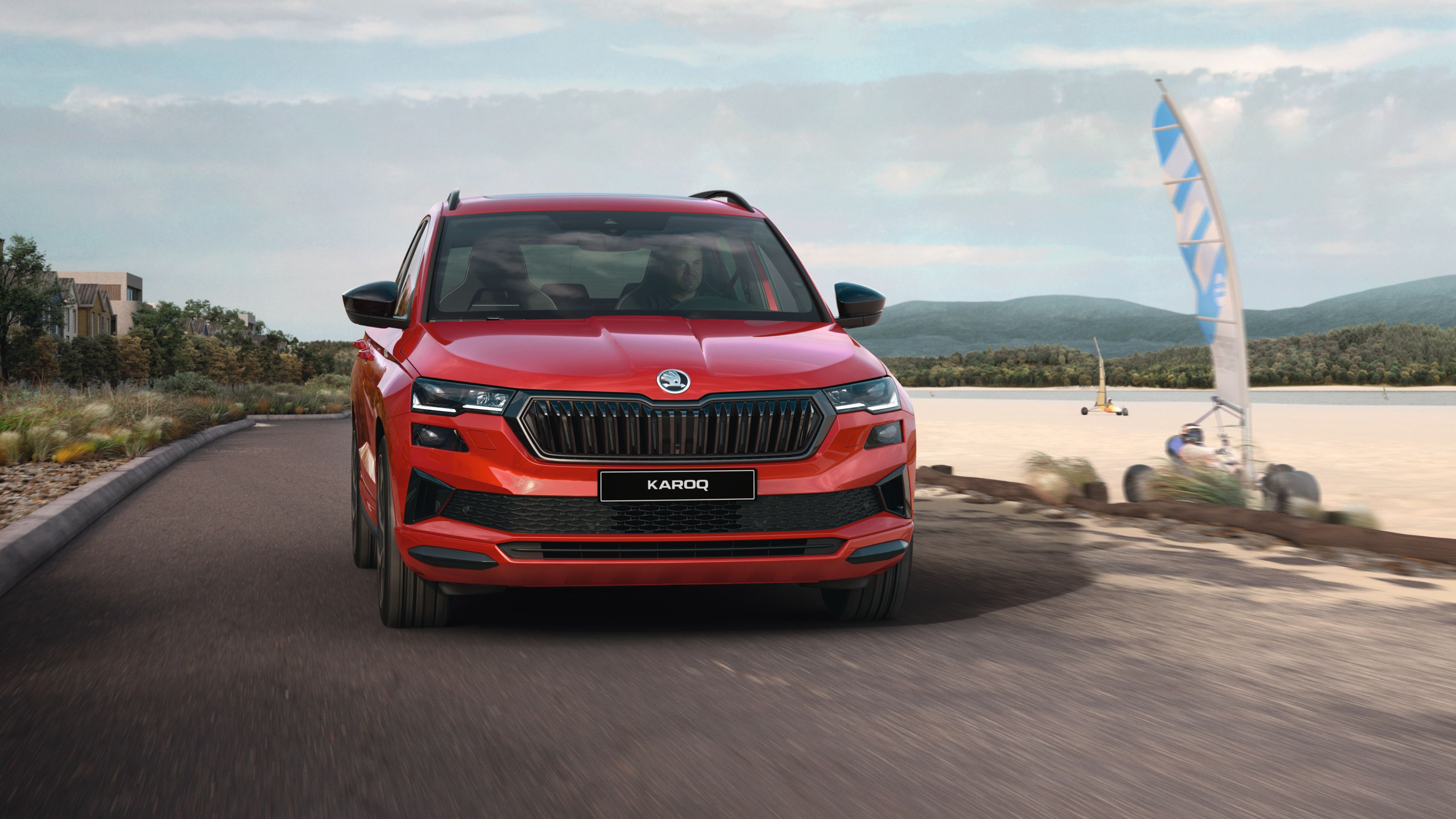 Rdeč SUV Škoda Karoq vozi po asfaltirani cesti ob obali, v ozadju pa so vidni morje, jadrnica in zgradbe.