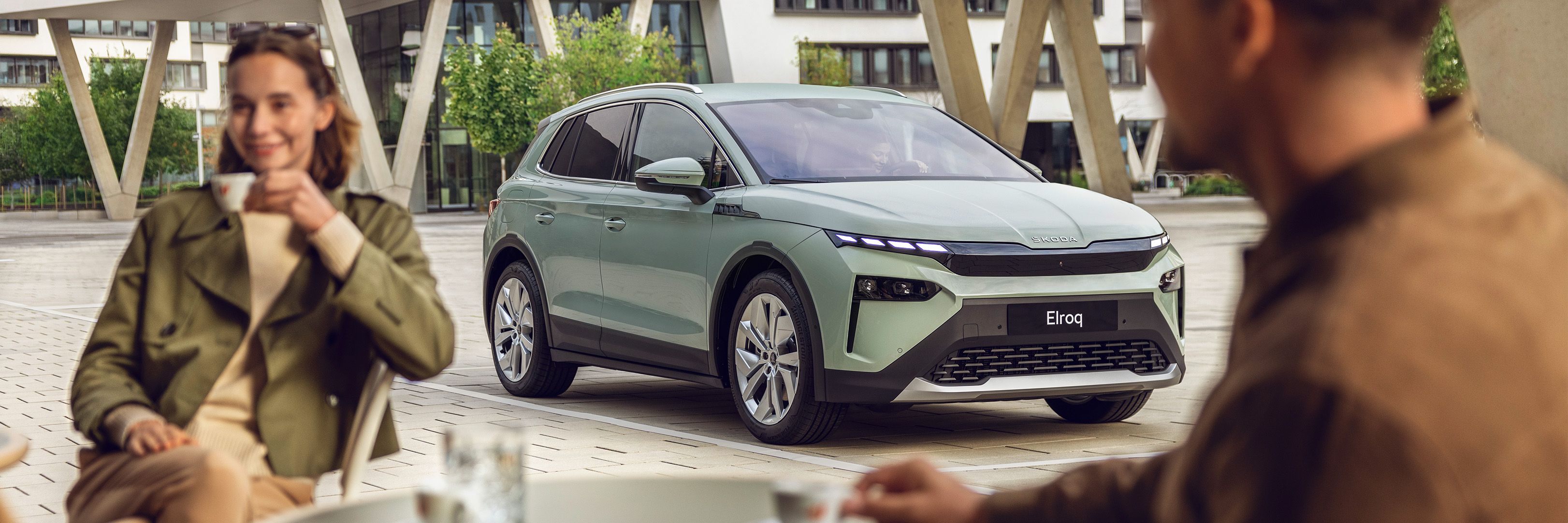 Električni avtomobil znamke Skoda, model Elroq, parkiran na prostem ob kavarni. Dva človeka sedita in pijeta kavo. Avto ima zelene odtenke in modernejši dizajn.