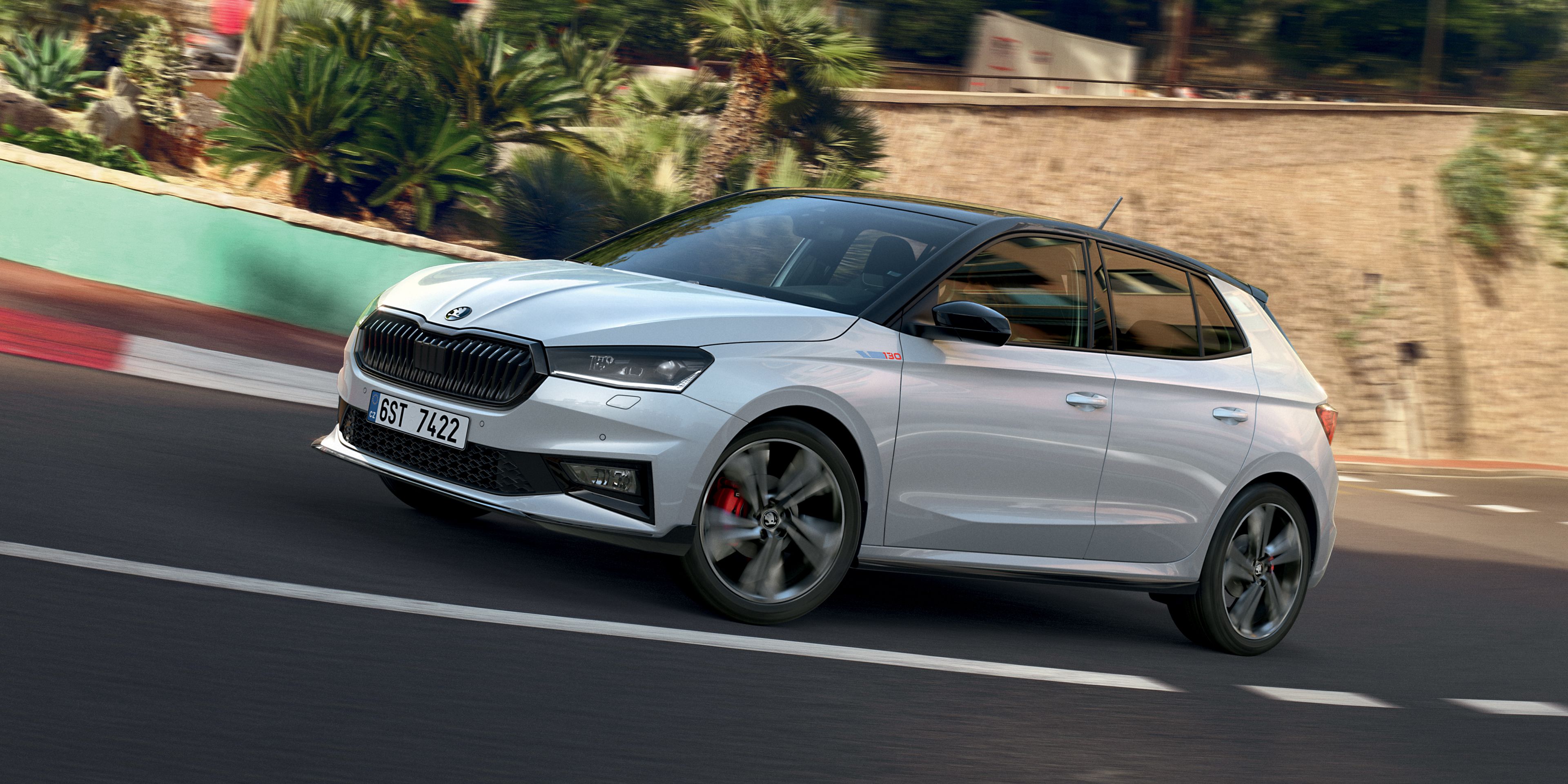 Srebrn športni avtomobil Škoda Fabia 130 vozi po cesti obdan z zelenjem. Avtomobil ima elegantno obliko in rdeče zavorne čeljusti. V ozadju je kamnita ograja in nekaj dreves.