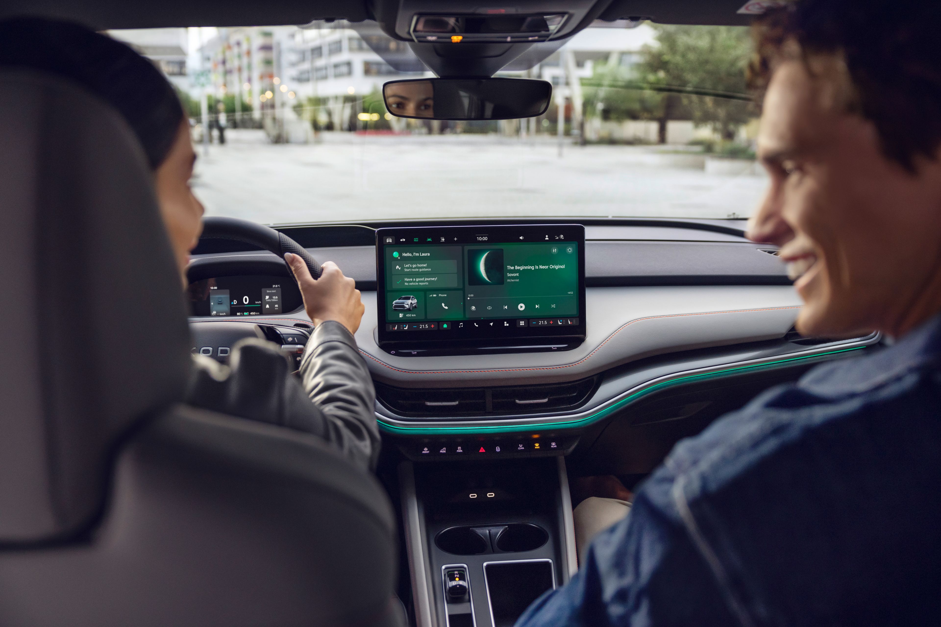 Dva človeka sedita v avtomobilu, eden za volanom. Armaturna plošča prikazuje sodoben zaslon na dotik z navigacijo in multimedijskimi informacijami. Okoli je urbana okolica s stavbami in zelenjem.