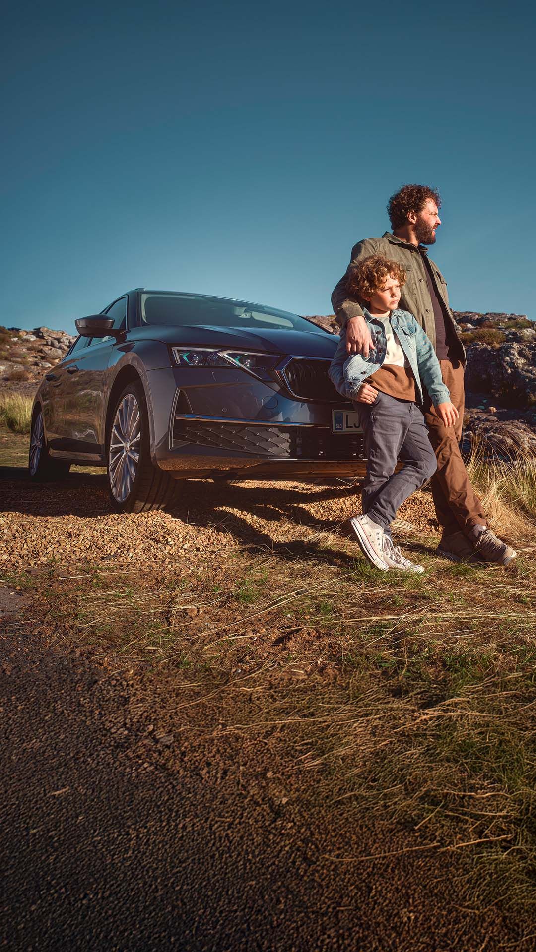 Moški stoji ob avtomobilu Škoda Octavia Combi na podeželski cesti, na travnatem območju. Oblečen je v rjavo jakno in hlače ter drži otroka, oblečenega v modro jakno in hlače. Ozadje je skalnato in travnato.