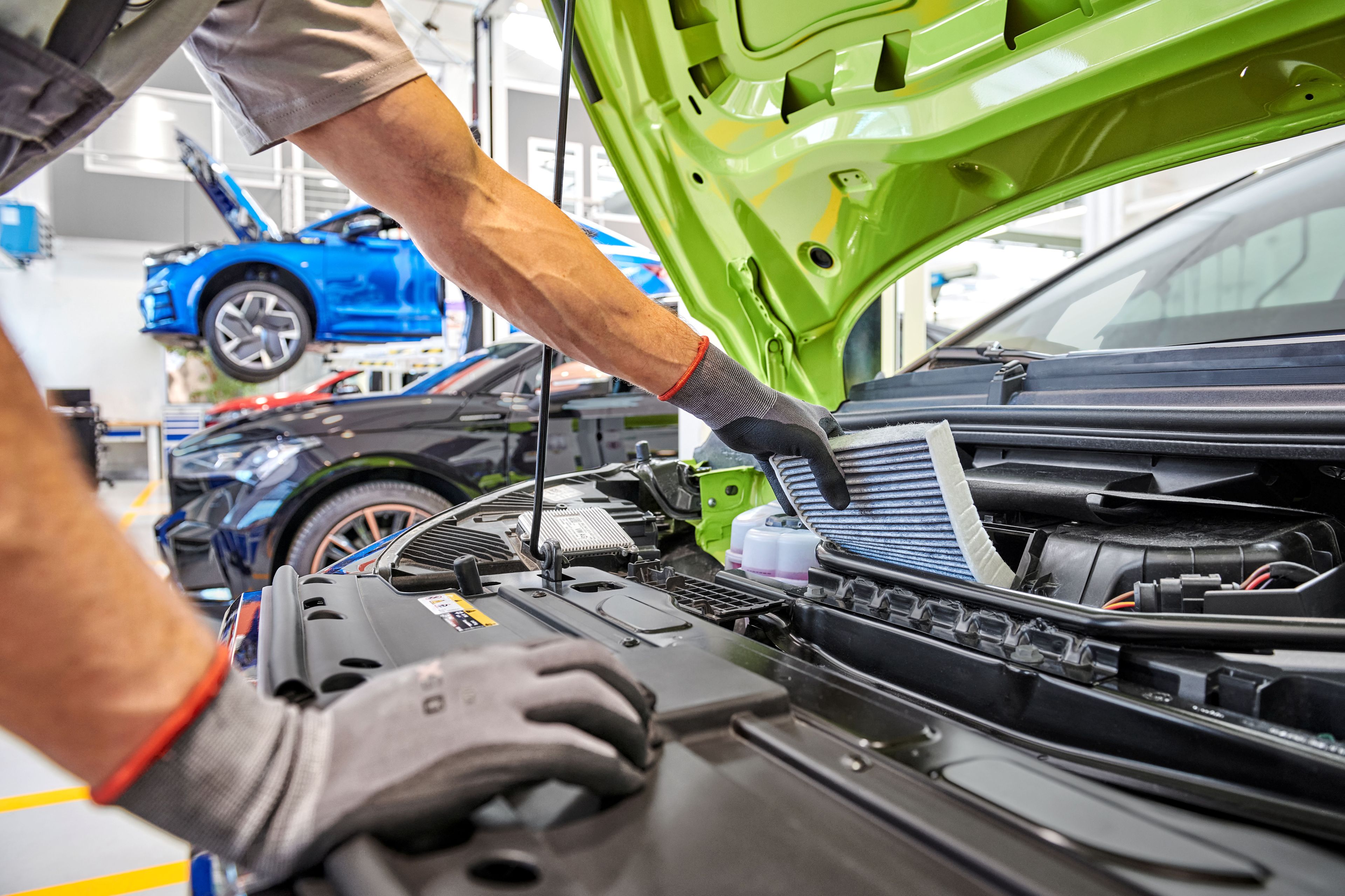 Mehanik med servisiranjem avtomobila v avtoservisu. Odprt motor avtomobila medtem ko mehanik pregleduje filter. V ozadju je več avtomobilov na dvigalu.