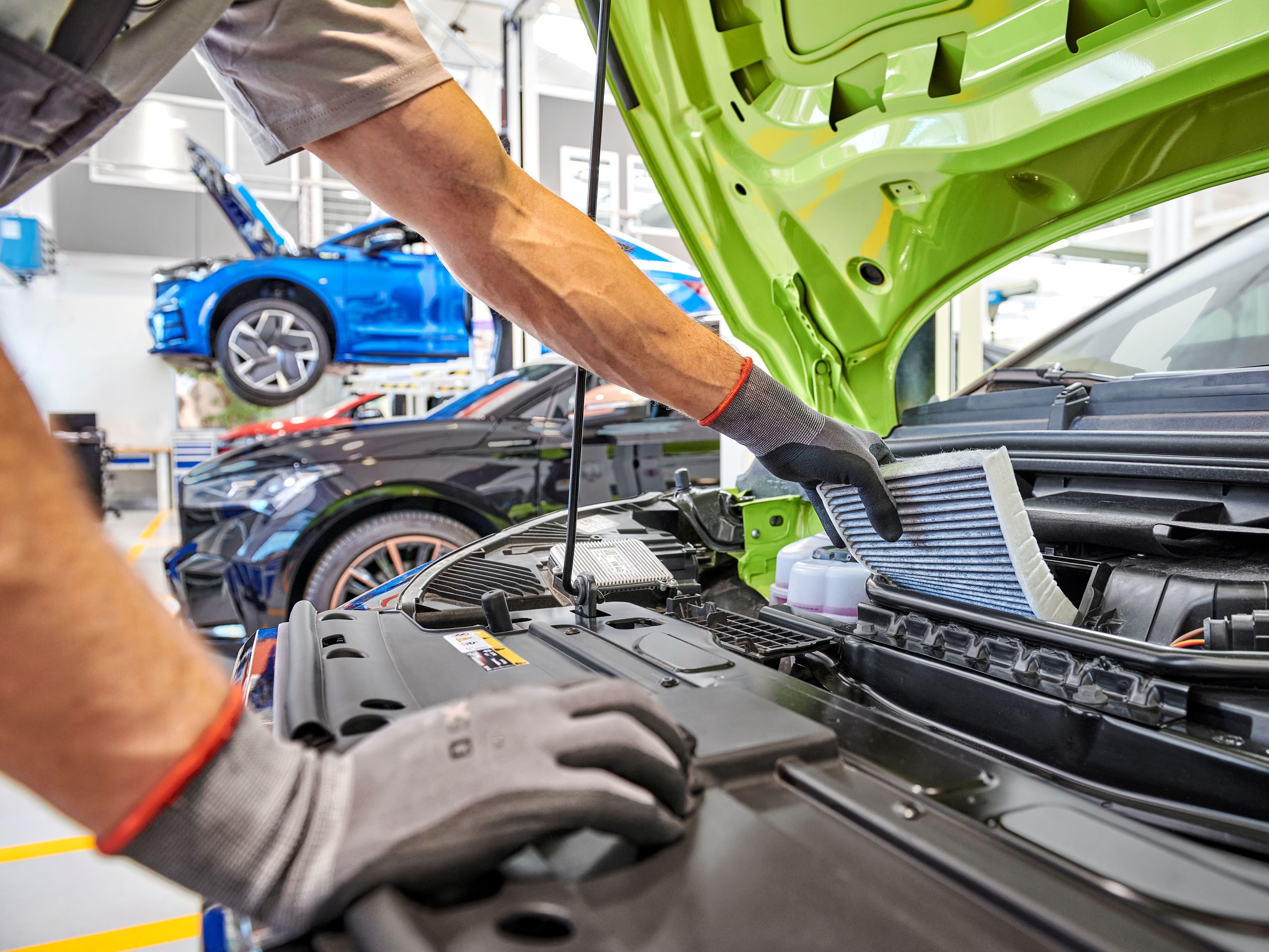 Mehanik med servisiranjem avtomobila v avtoservisu. Odprt motor avtomobila medtem ko mehanik pregleduje filter. V ozadju je več avtomobilov na dvigalu.