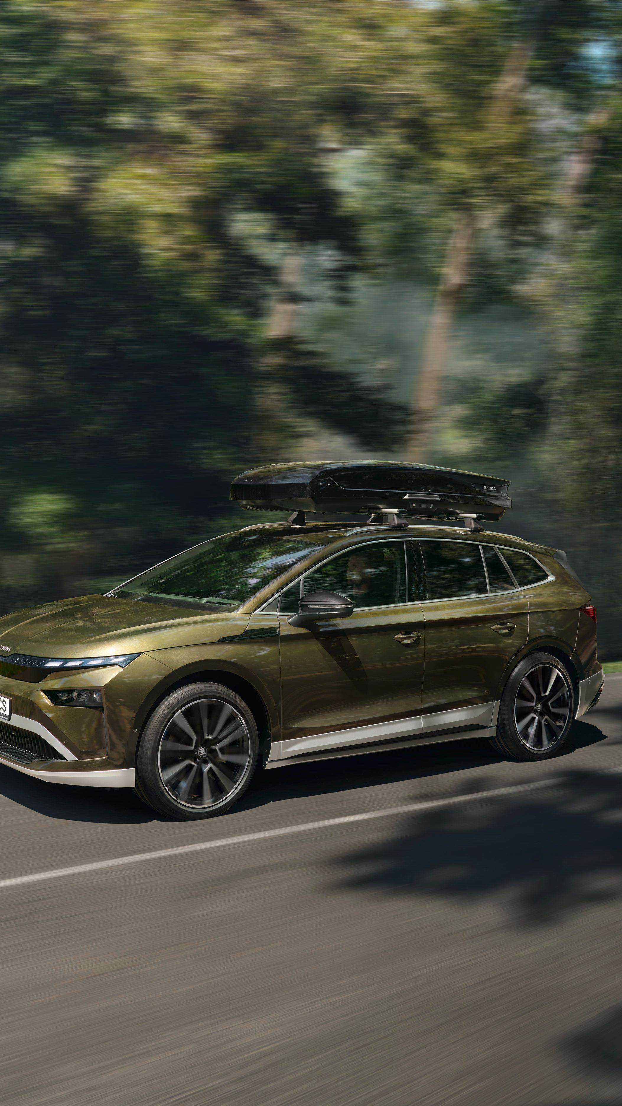 Avtomobil Kia EV9, ki vozi po cesti skozi gozdno območje. Vozilo je zeleno, aerodinamično oblikovano, na strehi ima pritrjeno strešno kovček. Cesta je ovita v bujno vegetacijo.