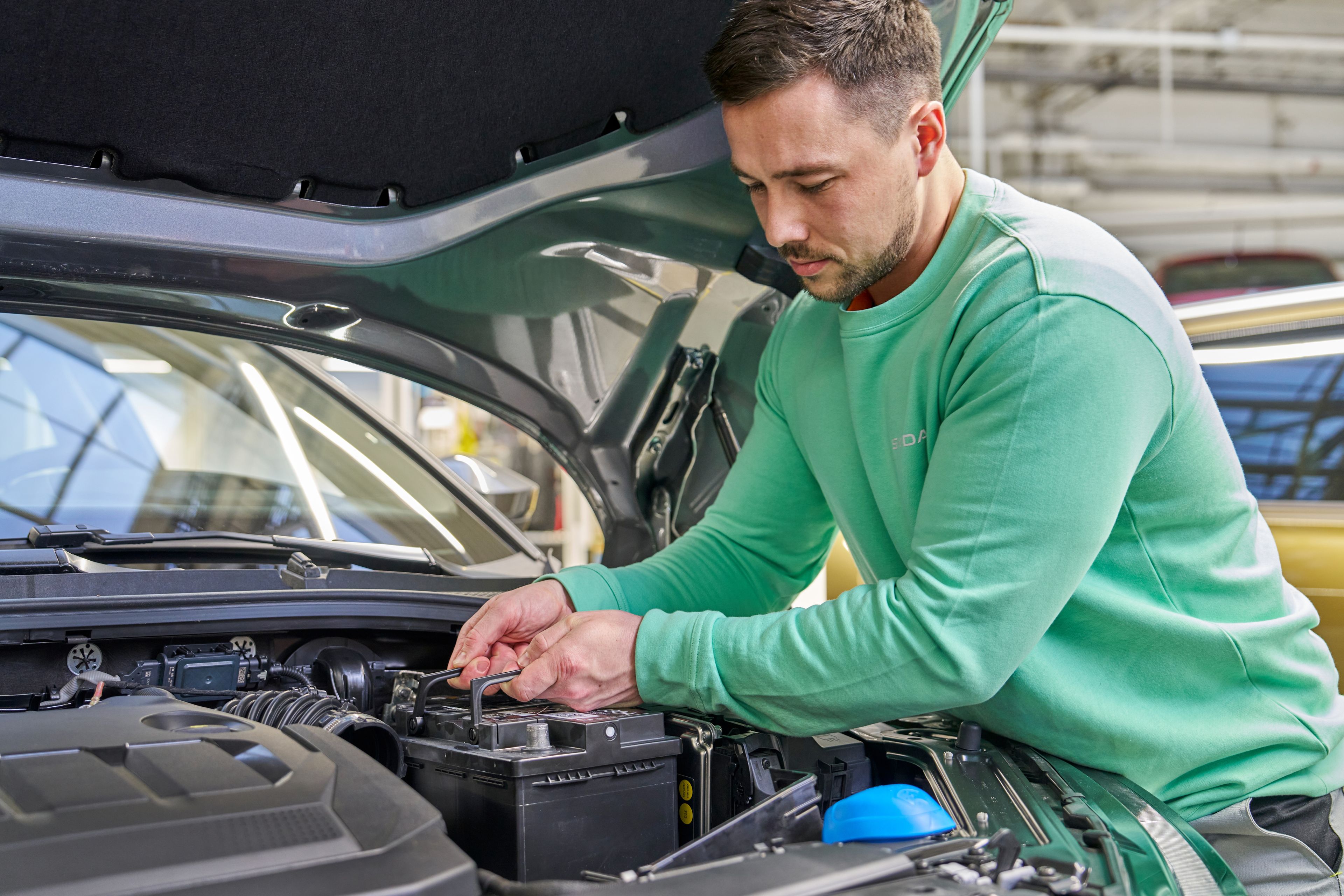 Oseba v zeleni majici je pod odprtim pokrovom avtomobila in dela z akumulatorjem. Pritrjuje ali popravlja akumulator.