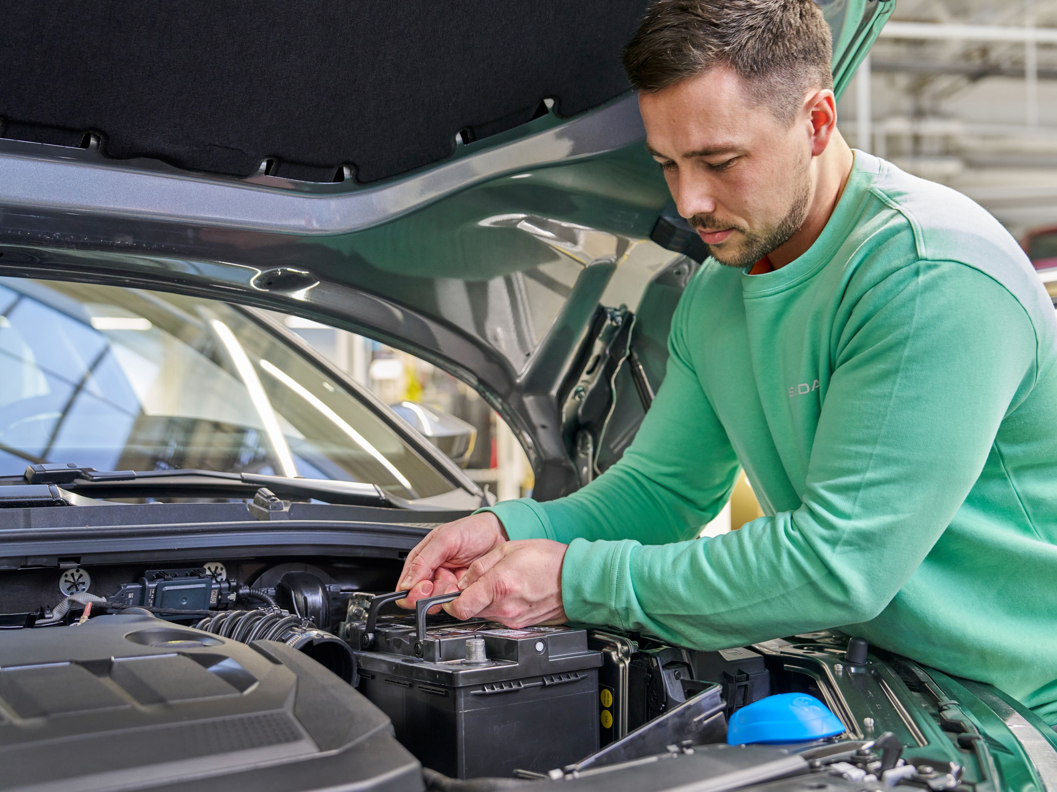 Oseba v zeleni majici je pod odprtim pokrovom avtomobila in dela z akumulatorjem. Pritrjuje ali popravlja akumulator.