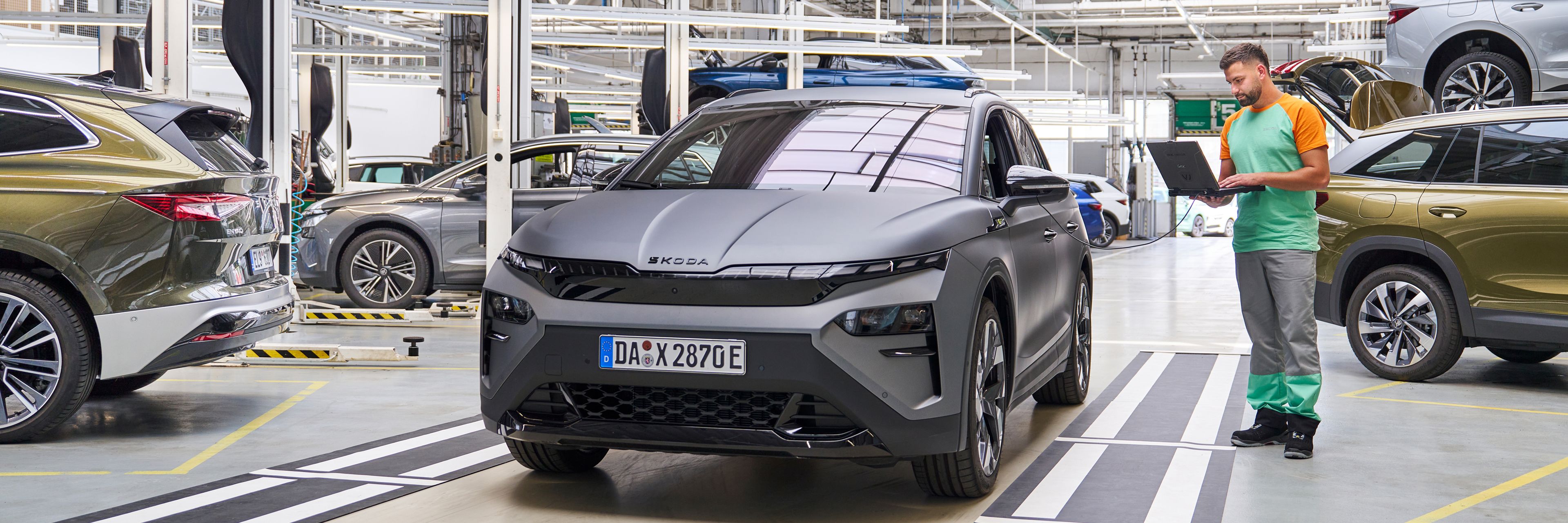 Siva avtomobil stoji v delavnici, medtem ko oseba v delovni uniformi zapiše nekaj v beležko. V ozadju so še drugi avtomobili na dvigalih, ki se očitno servisirajo.