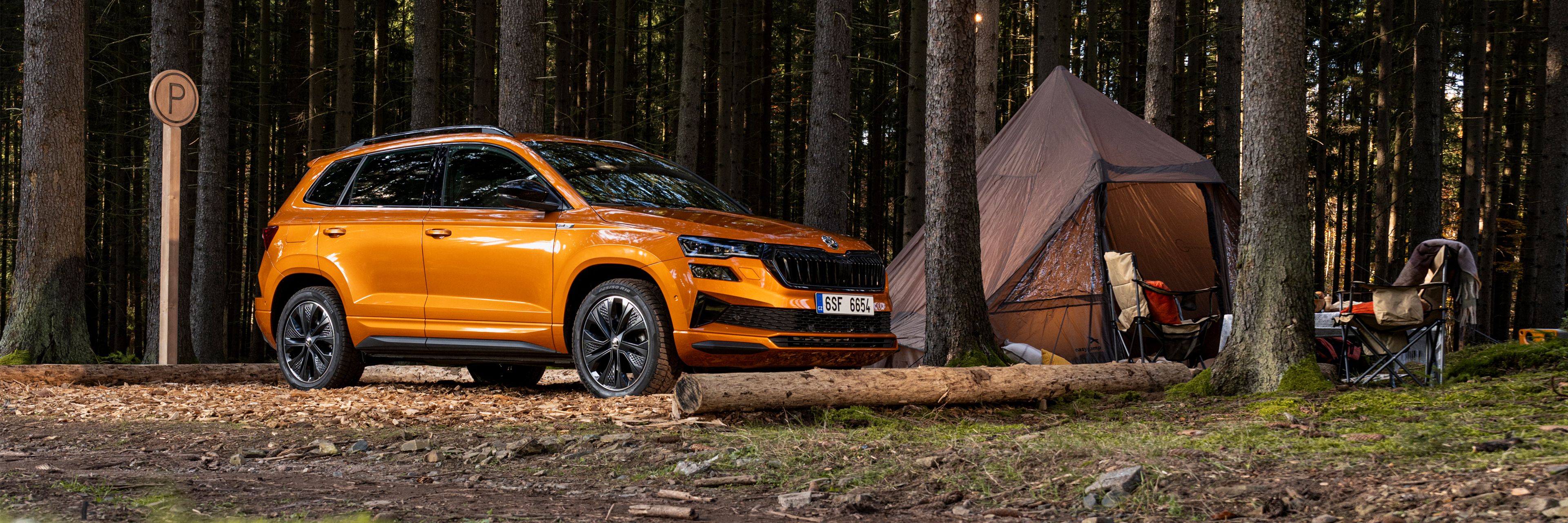 Ornažen SUV, Škoda Karoq, je parkiran ob kampu v gozdu. Avto je obdan z visokimi drevesi, zraven pa stoji šotor in zložljiv stol. V ozadju so vidne lučke, ki dajejo občutek topline.