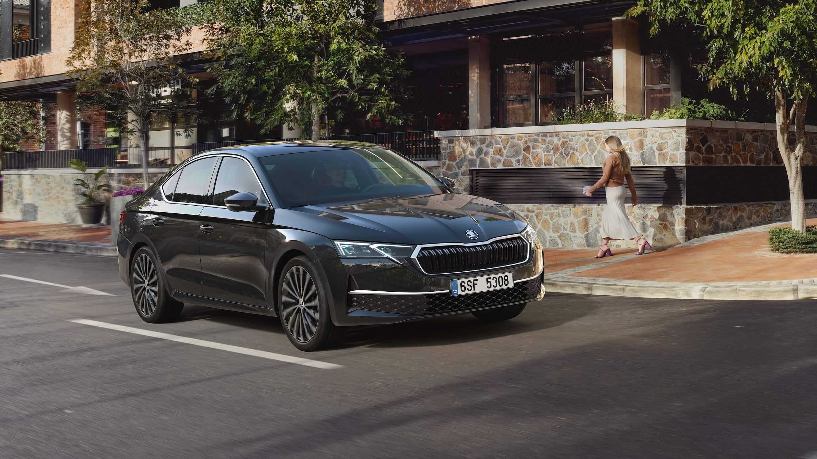 Črna limuzina Škoda Octavia vozi po mestni ulici. V ozadju so zgradbe, drevesa in sprehajalka. Avto je parkiran s številko registrske tablice "6SF 5308".
