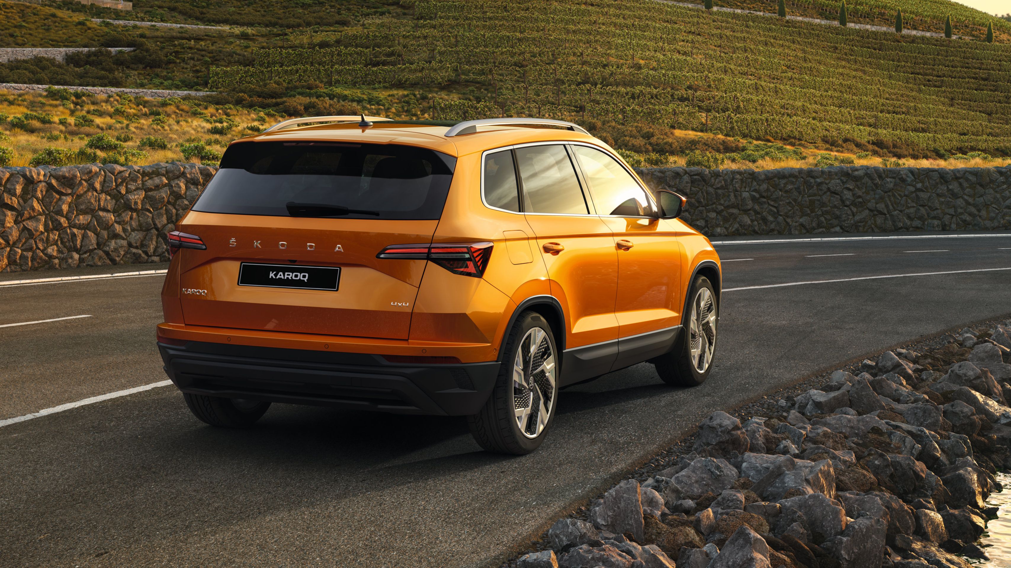 Oranžni Škoda Karoq parkiran ob podeželski cesti z kamnito ograjo in zelenimi polji v ozadju.