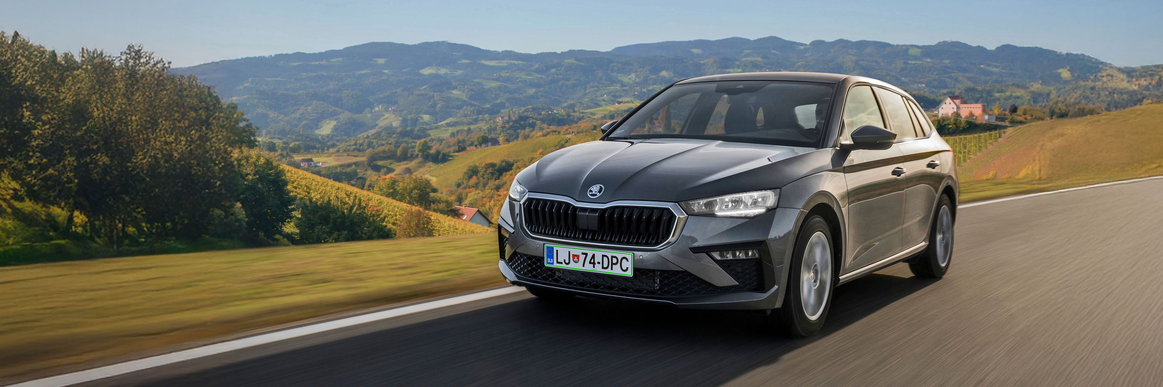 Siva limuzina Škoda Scala , ki vozi po podeželski cesti, obdani z griči in vinogradi. Na registrski tablici je oznaka "LJ". Ne bo izpostavljati nekaj specifičnega.