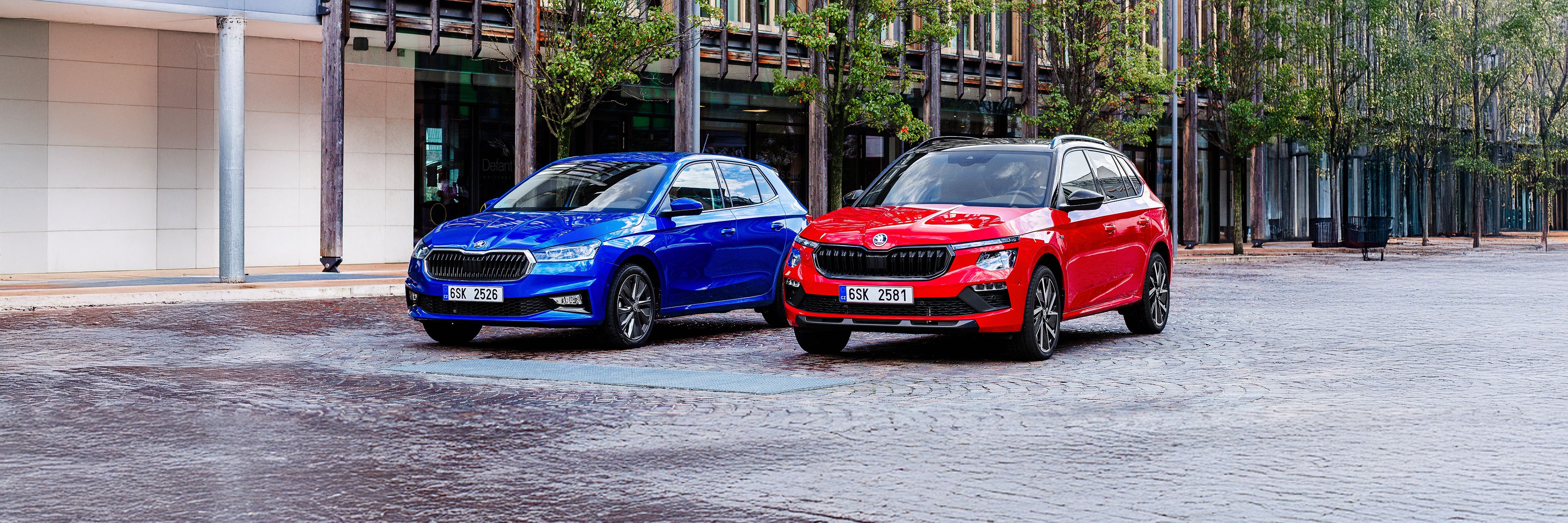 Na sliki sta rdeči Škoda Kamiq in modra Škoda Scala, parkirana na tlakovani površini pred modernimi zgradbami. Levo je modri avto, desno pa rdeči. V ozadju so drevesa in steklene fasade.