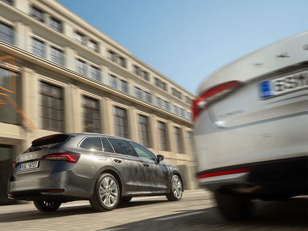 Dva avtomobila se vozita mimo velike sodobne stavbe z modrim nebom v ozadju. Tlakovana cesta je jasna in ima vzorec. Avtomobil v ospredju je delno zamegljen, medtem ko je avto v ozadju bolj jasen.