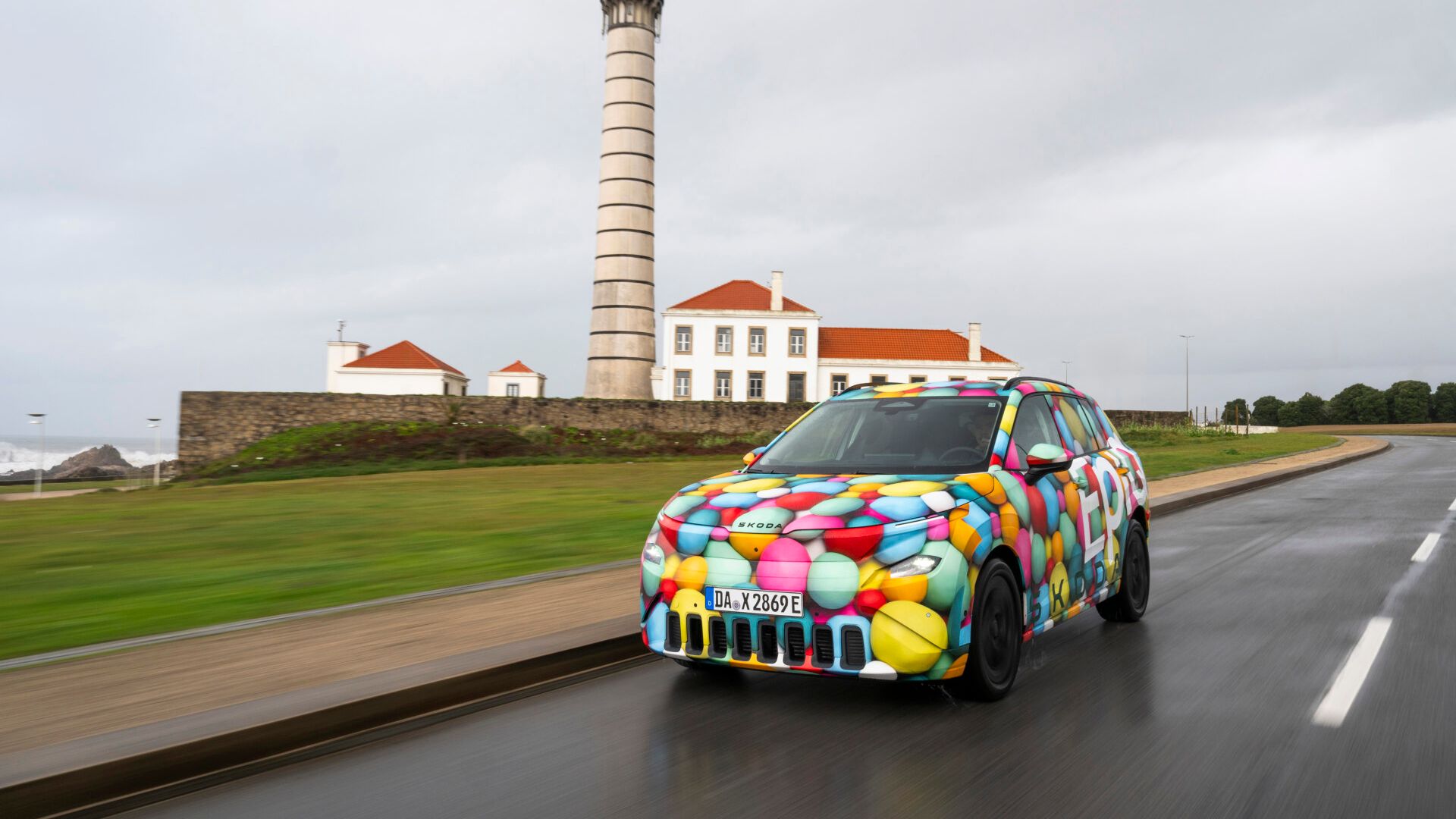 Avtomobil, Škoda Epiq, živih barv pelje po cesti ob obali z visokim svetilnikom in stavbo v ozadju. Nebo je oblačno in cesta mokra.