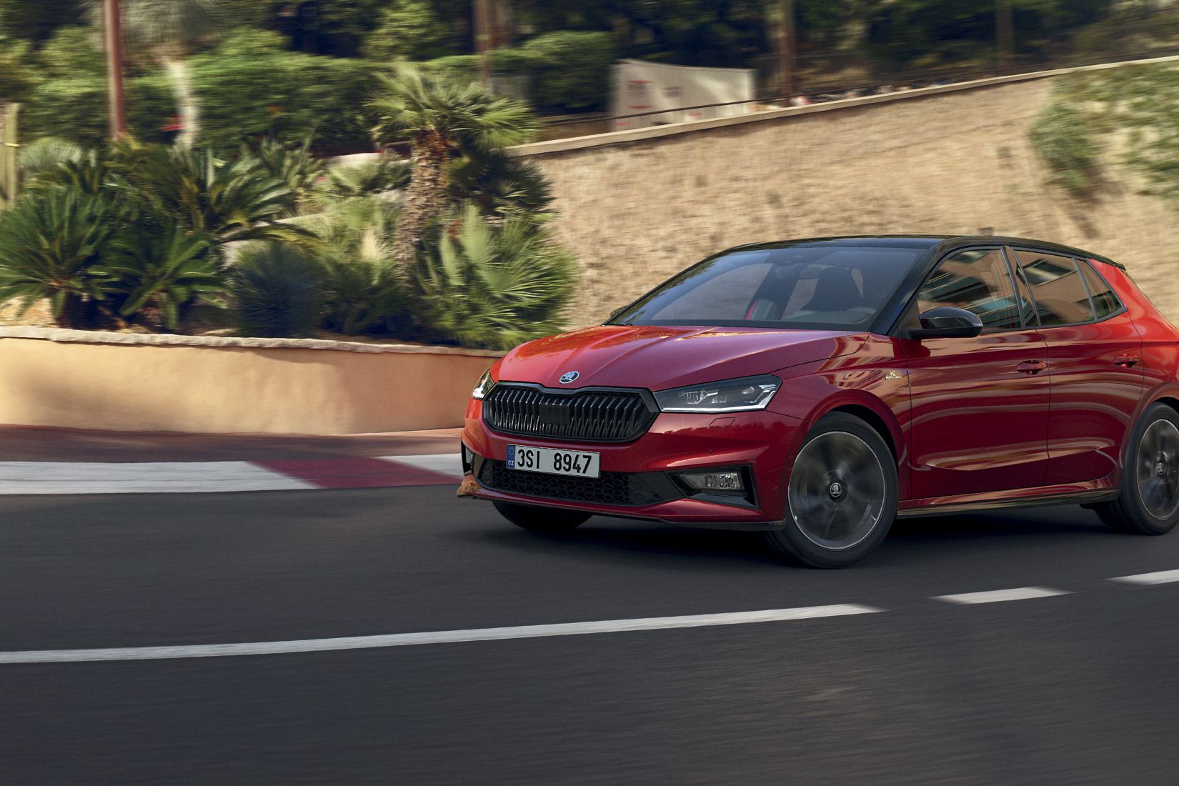 Rdeča Fabia Monte Carlo avtomobil se vozi po vijugasti cesti, obdani z zelenjem in kamnitimi zidovi. V ozadju je motociklist. Scena je postavljena v sončnem vremenu.