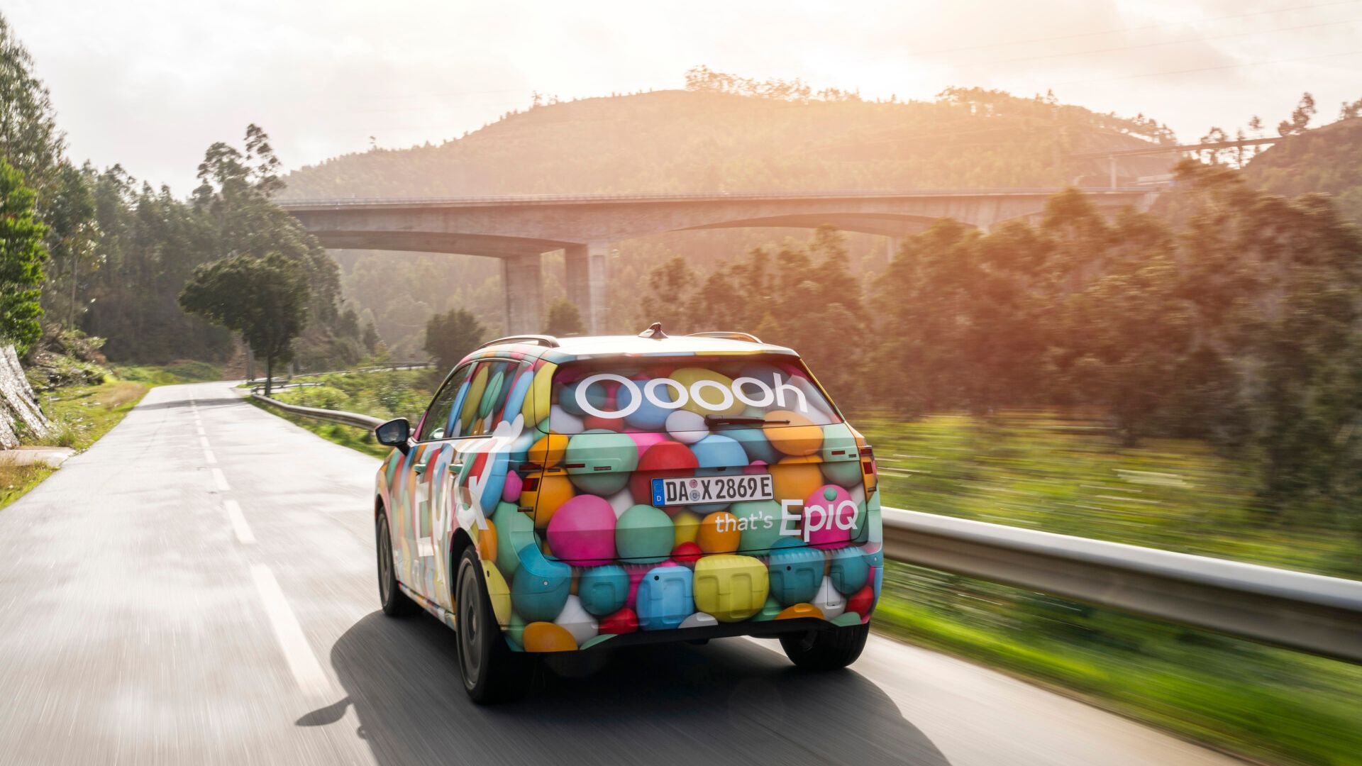 Škoda Epiq z živopisno grafiko na zadnjem delu, ki vsebuje barvne krogle in besede "Oooh that’s Epic", vozi po podeželski cesti obdan z zelenjem in hribi. Cesta vodi pod velik most, in vreme je sončno.