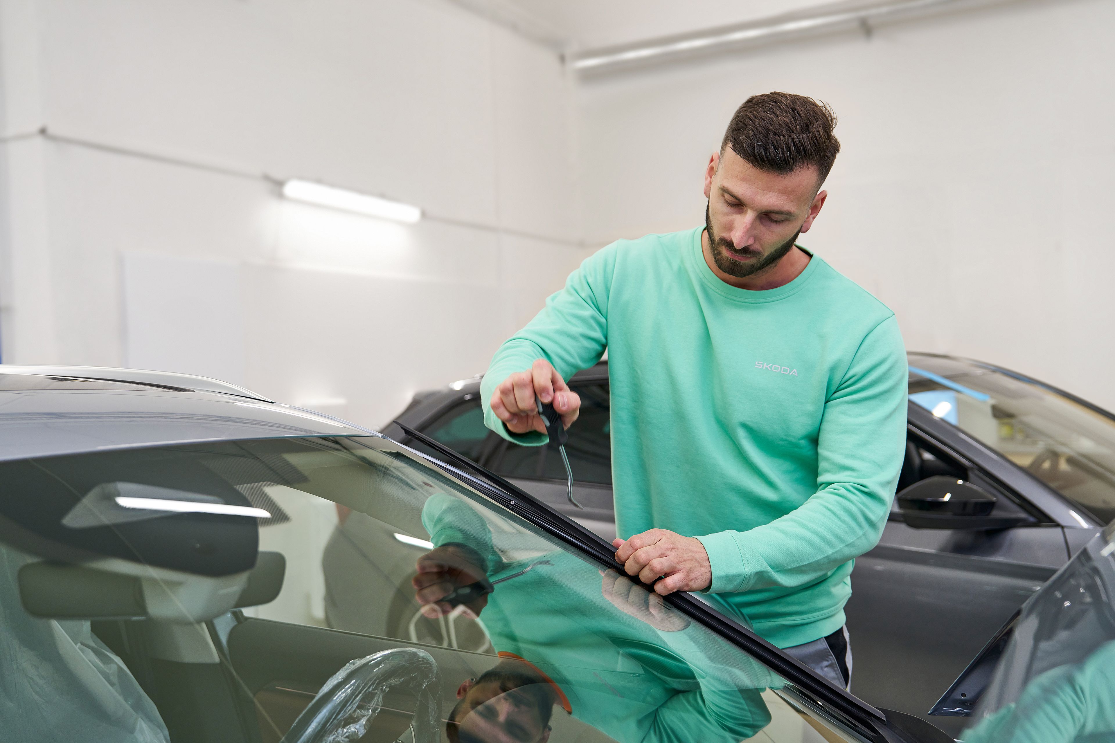 Človek v svetlozeleni majici se ukvarja s popravilom avtomobilskega vetrobranskega stekla v delavnici.