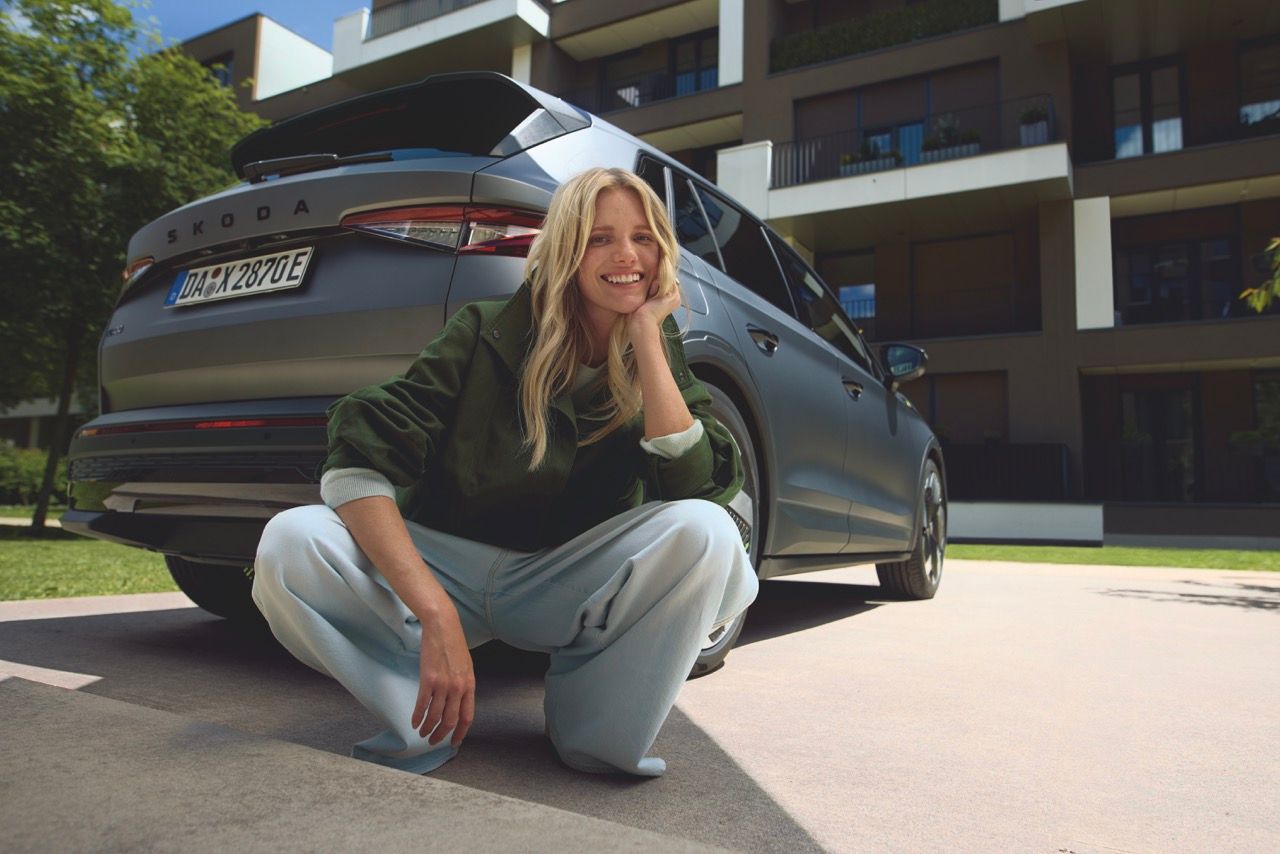 Oseba v priloženih hlačah počepne pred parkiranim avtomobilom znamke Škoda. V ozadju je moderna večstanovanjska stavba.