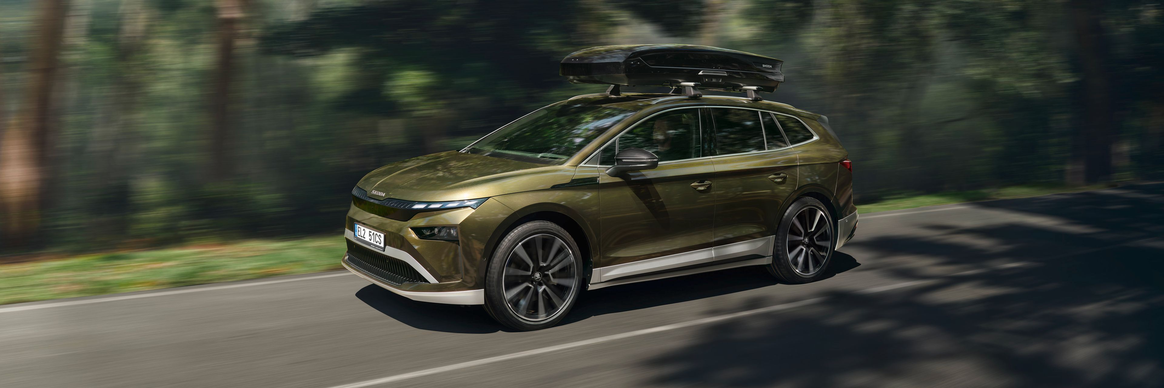 Avtomobil Kia EV9, ki vozi po cesti skozi gozdno območje. Vozilo je zeleno, aerodinamično oblikovano, na strehi ima pritrjeno strešno kovček. Cesta je ovita v bujno vegetacijo.