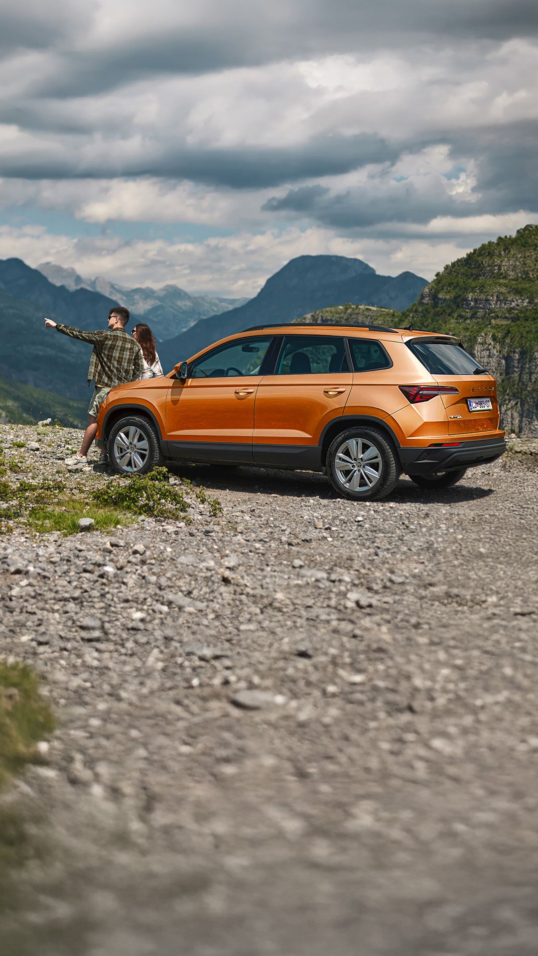 Oranžni SUV Škoda Karoq je parkiran na kamnitem parkirišču v goratem okolju. Oseba z zabrisanim obrazom kaže na oddaljeno točko v pokrajini. V ozadju so vidni visoki hribi in oblačno nebo.