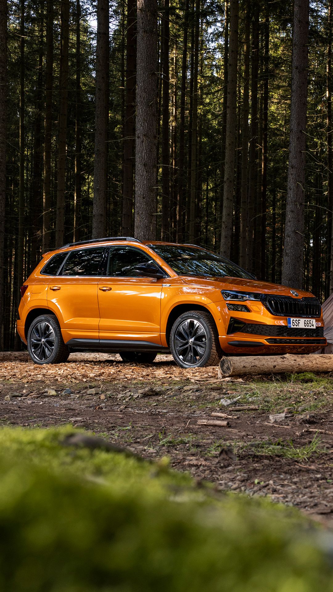 Svetlo oranžen SUV Škoda Karoq je parkiran v gozdu med visokimi drevesi. Čista in moderna oblika vozila izstopa v naravni okolici. Na spodnjem delu registrske tablice so črne številke na belem ozadju.