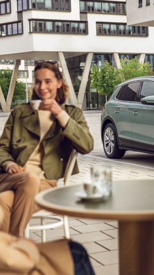 Na sliki je sodoben električni avtomobil, parkiran na mestnem trgu. Dva človeka sedita za mizo in pijeta kavo ter opazujeta avtomobil. Ozadje prikazuje večnadstropno stavbo s steklenimi okni.