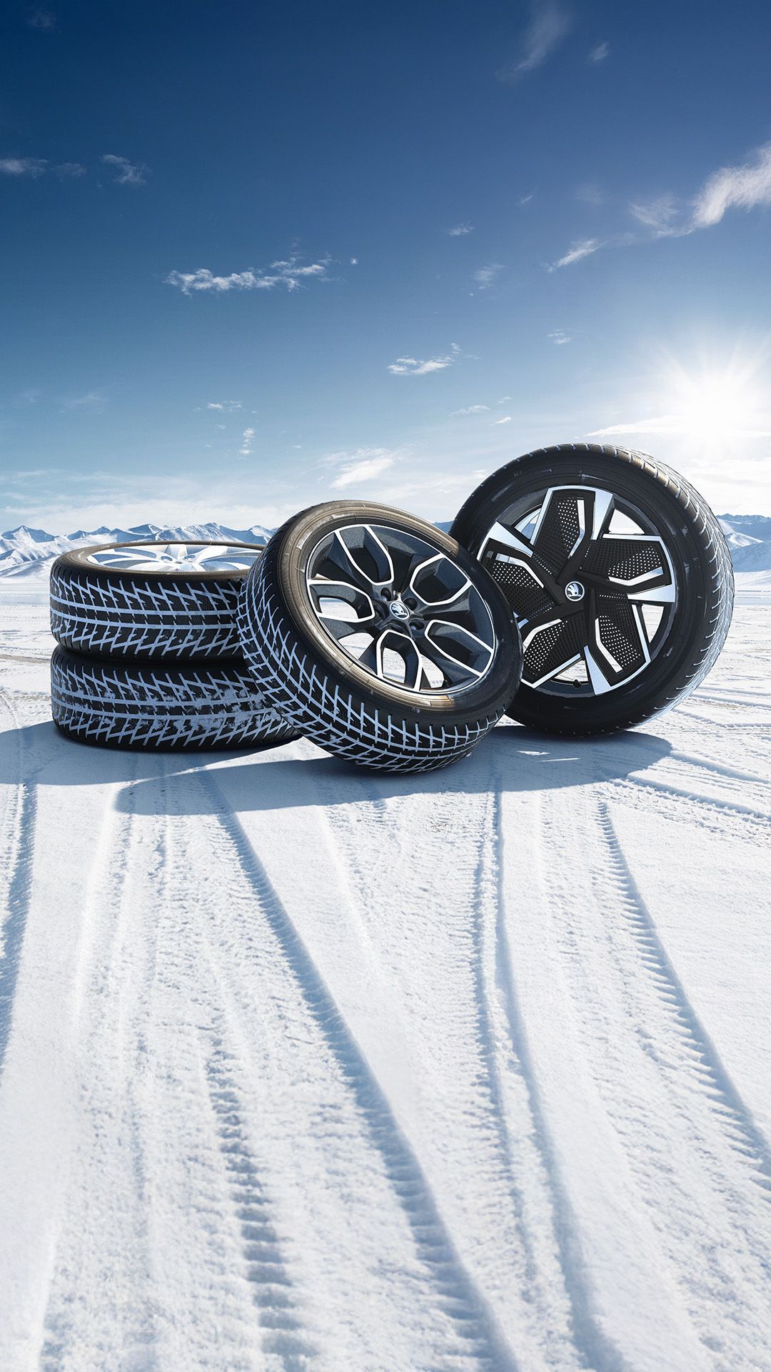 Slika prikazuje štiri avtomobilske pnevmatike na snegu ob sončnem dnevu. V ozadju so vidne zasnežene gore.