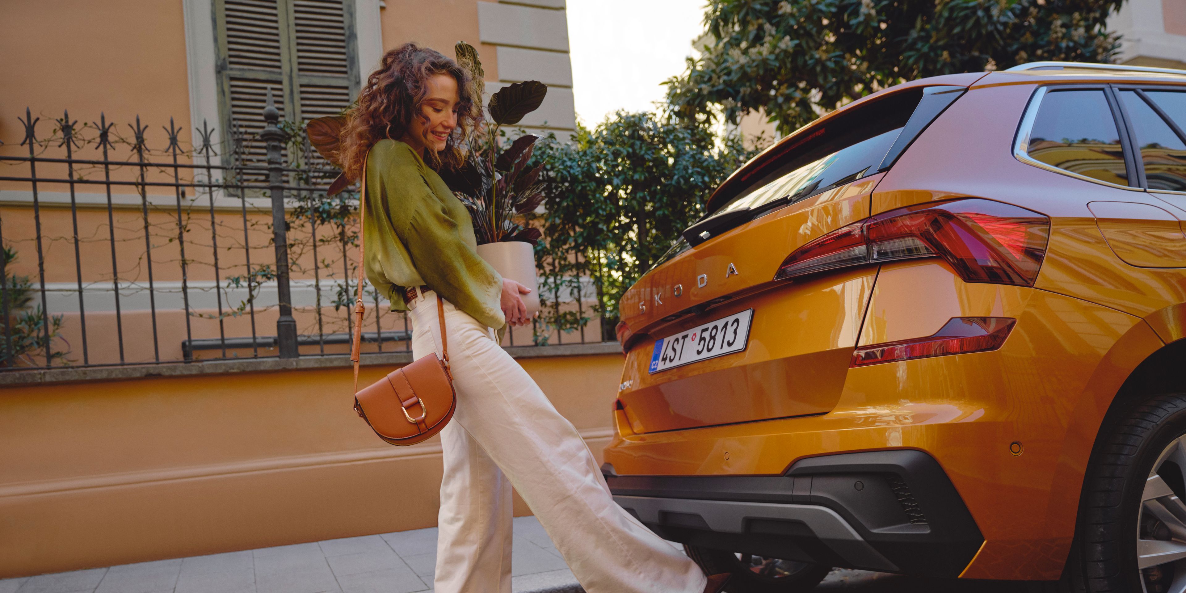 Oseba stoji ob oranžnem vozilu, Škoda Kamiq, parkiranem na ulici pred stavbo. Ima oblečene bele hlače in zeleno bluzo ter nosi rjavo torbico čez ramo. V rokah drži večjo rastlino v lončku.