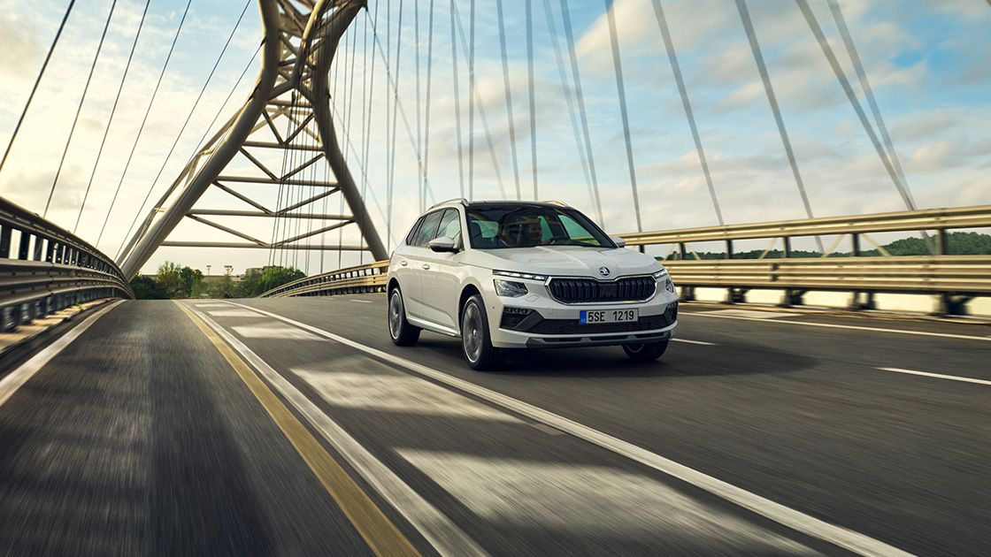 Avtomobil znamke Škoda vozi po modernem mostu ob sončnem dnevu. Vozilo je bele barve in ima močnejše linije. V ozadju so vidna jeklena konstrukcija mostu in narava.