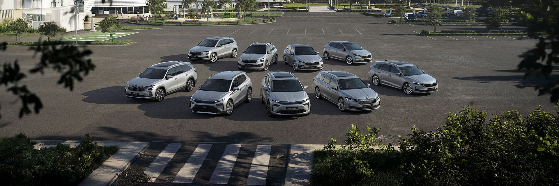 Na velikem parkirišču stoji 10 avtomobilov znamke Škoda, razporejenih v dve vrsti. V ozadju sta vidni moderna stavba in več stanovanjskih objektov. Nebo je jasno in modro, nekaj dreves pa obdaja parkirišče.