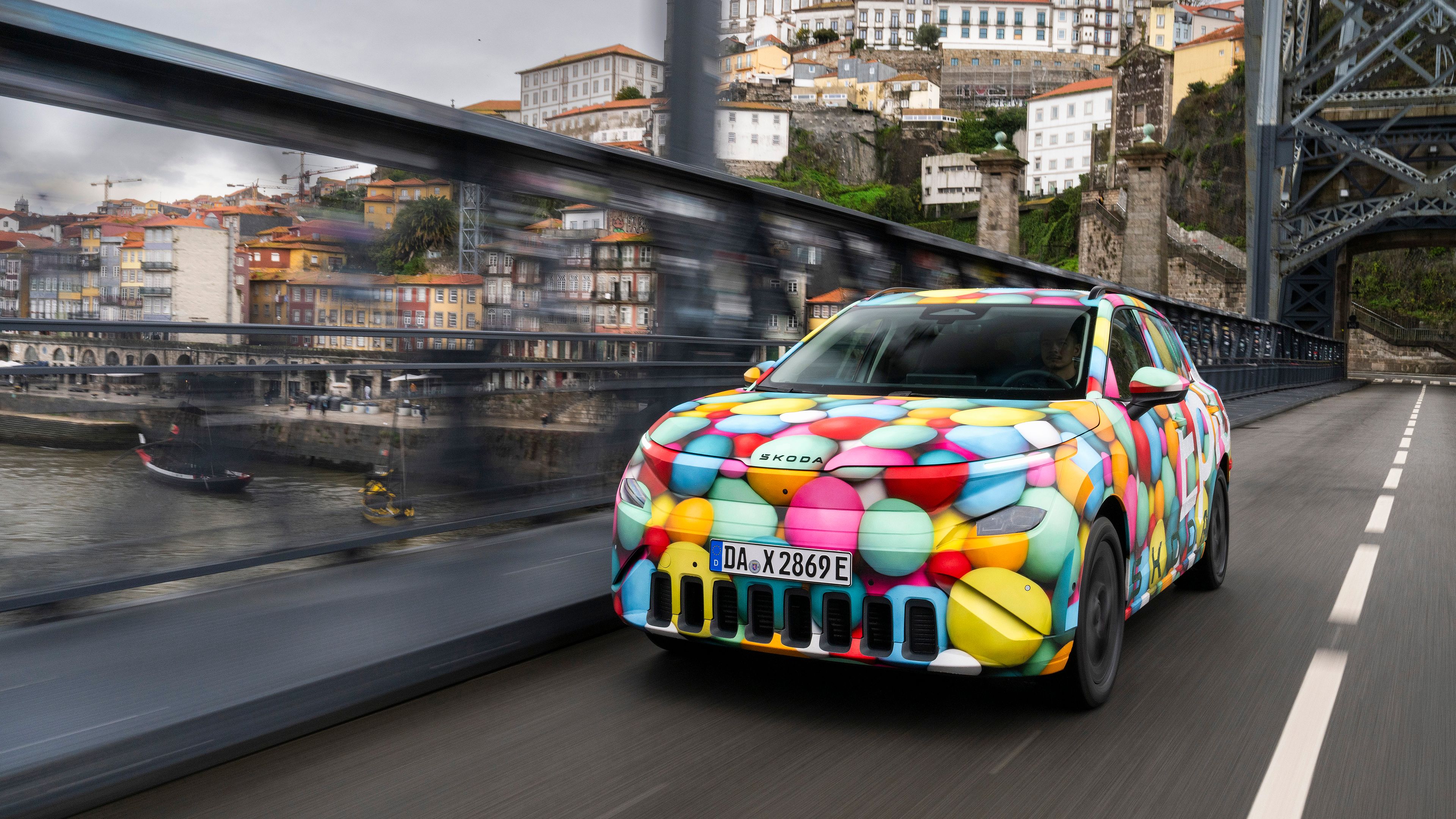 Vozilo Škod Epiq s pisanimi vzorci pelje po mostu čez reko v urbano okolje z več stavbami na hribovitem območju. Nebo je oblačno.