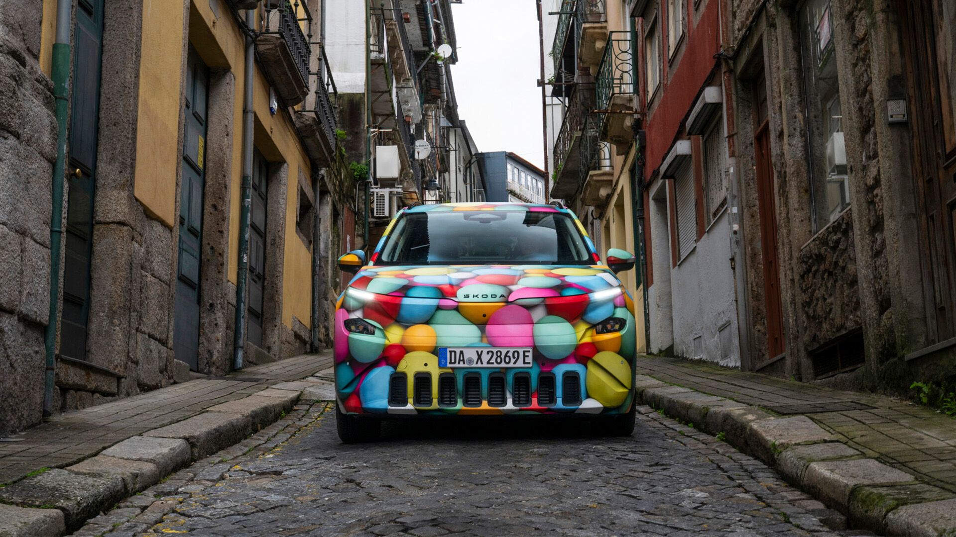 Škoda Epiq, prekrit s pisanimi kroglicami, je parkiran na kamniti cesti med starimi stavbami v ozki ulici.