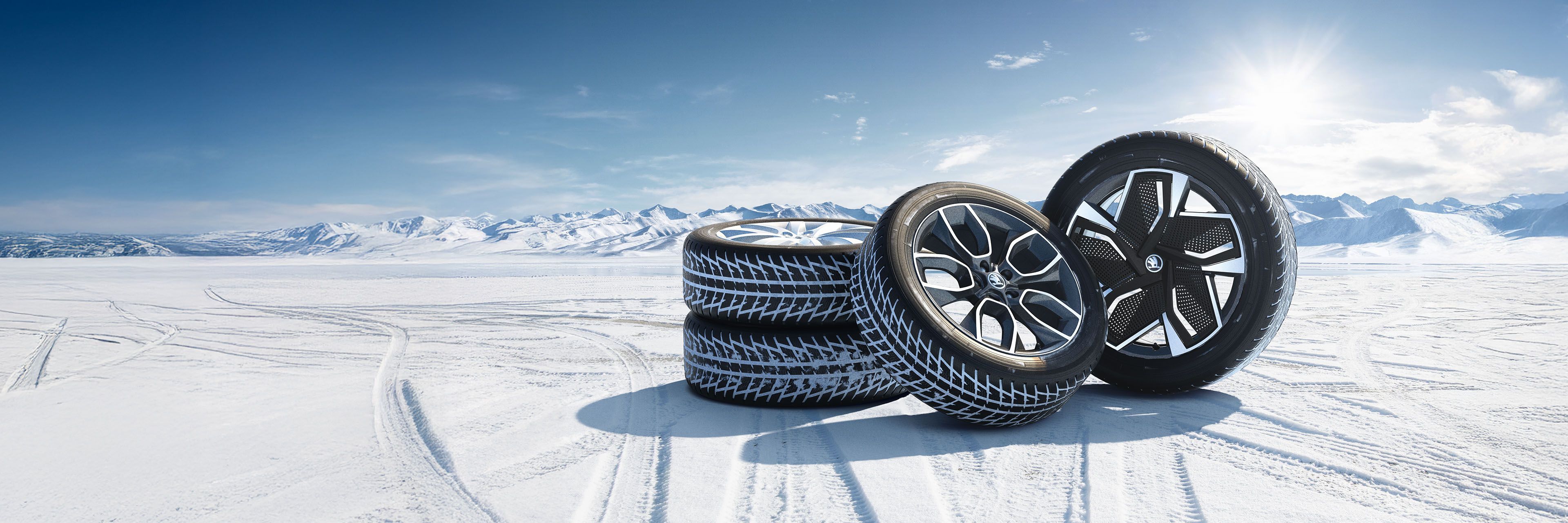 Štiri zimske gume so postavljene na snežno pokrajino z zasneženimi gorami v ozadju.
