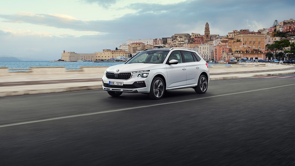 Bel avtomobil Škoda Kamiq se vozi po cesti ob obali, z mestom in morjem v ozadju. Nebo je delno oblačno.