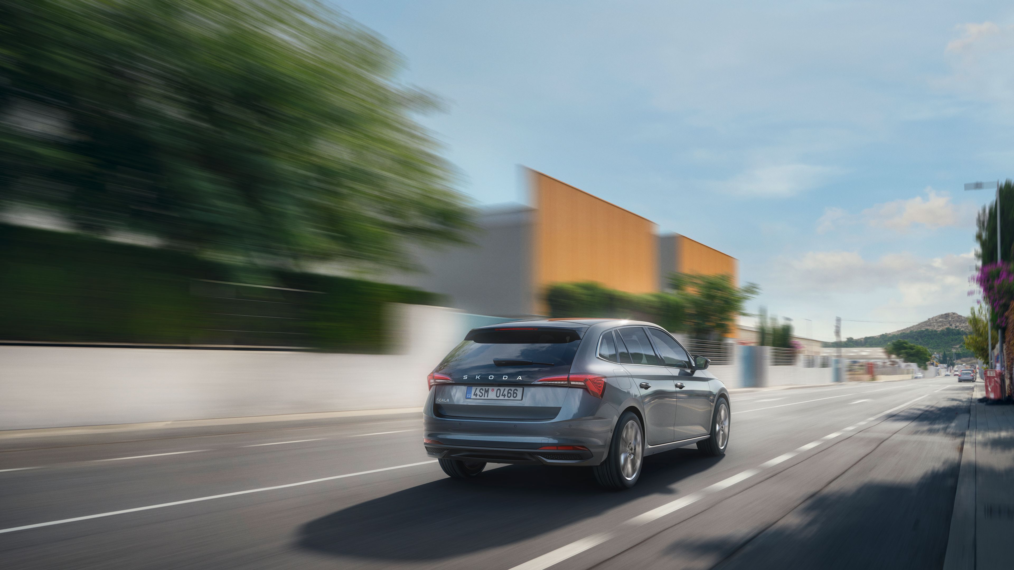 Na sliki je siv osebni avtomobil, ki se hitro pelje po mestni ulici. Ulica je obdana z modernimi zgradbami in drevesi. Njegova registrska tablica je vidna.