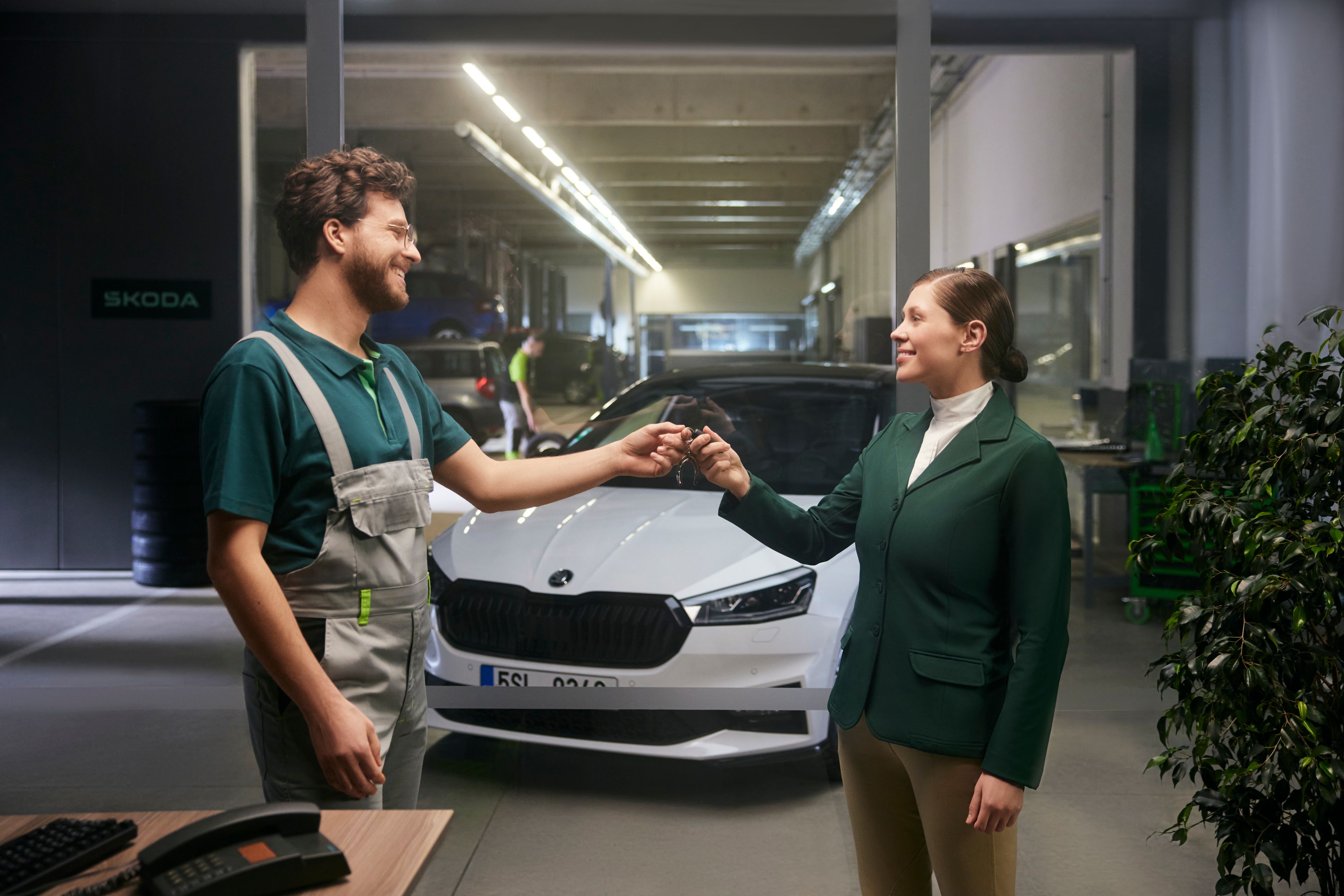 Moški in ženska stojita v mehanični delavnici in si izmenjujeta ključe avtomobila.