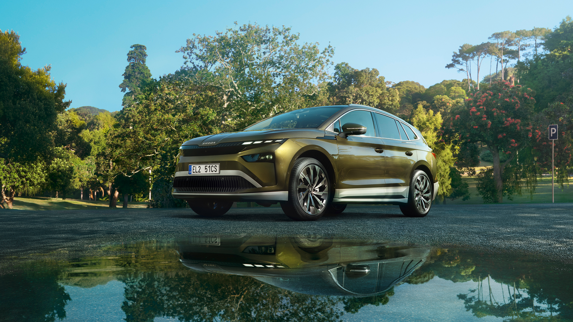 Električni avto, Škoda Enyaq, parkiran na asfaltni površini sredi zelenega parka, obdan z drevesi in grmičevjem. V ozadju je jasno modro nebo. Spodnja prednja stran avtomobila se vidi v odsevu luže na tleh.