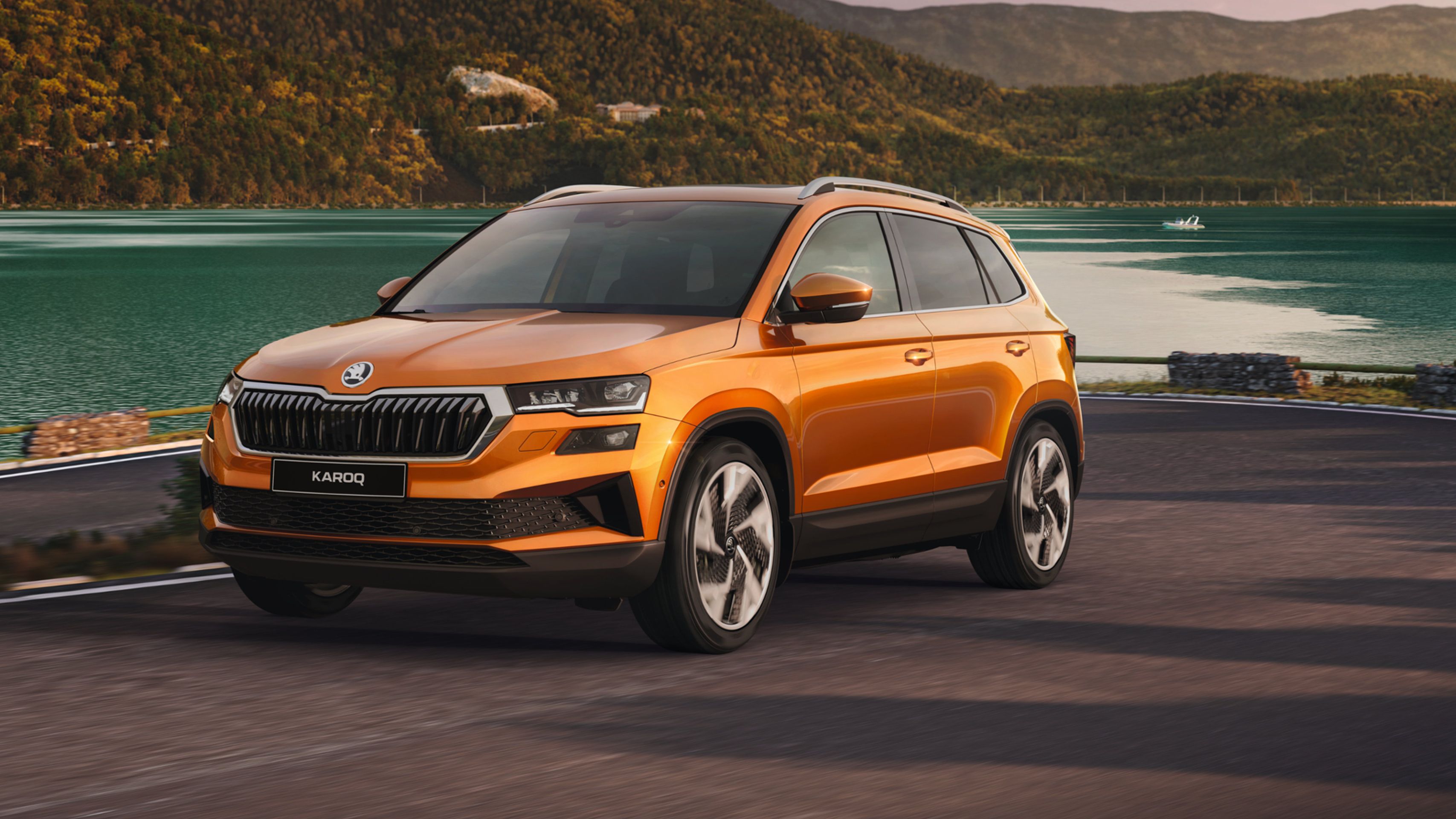 Oranžni SUV vozilo Škoda Karoq parkiran ob velikem jezeru z zelenimi bližnjimi hribi v ozadju. Vozilo ima moderne linije, velika kolesa in odprto panoramsko streho. V ozadju je tudi lesena ograja ter kamnita pot.