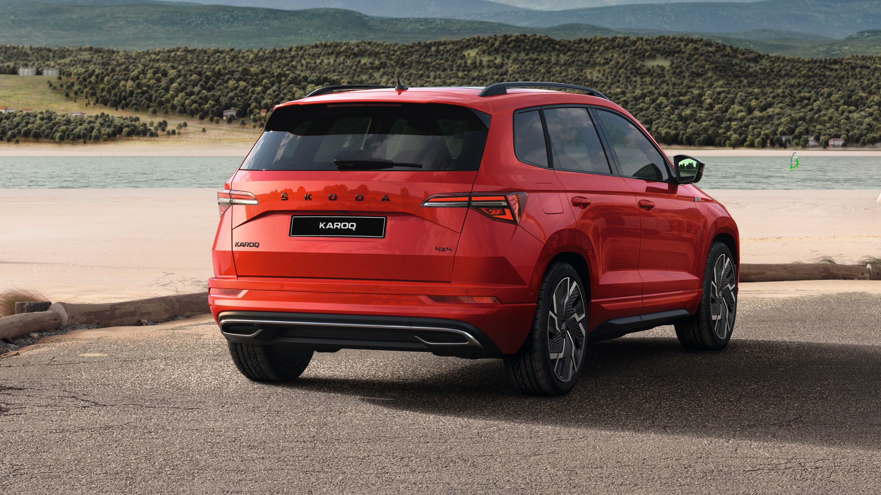 Rdeč športni terenec Škoda Karoq parkiran ob obali, v ozadju pa oseba na majhni jadrnici ob robu jezera.