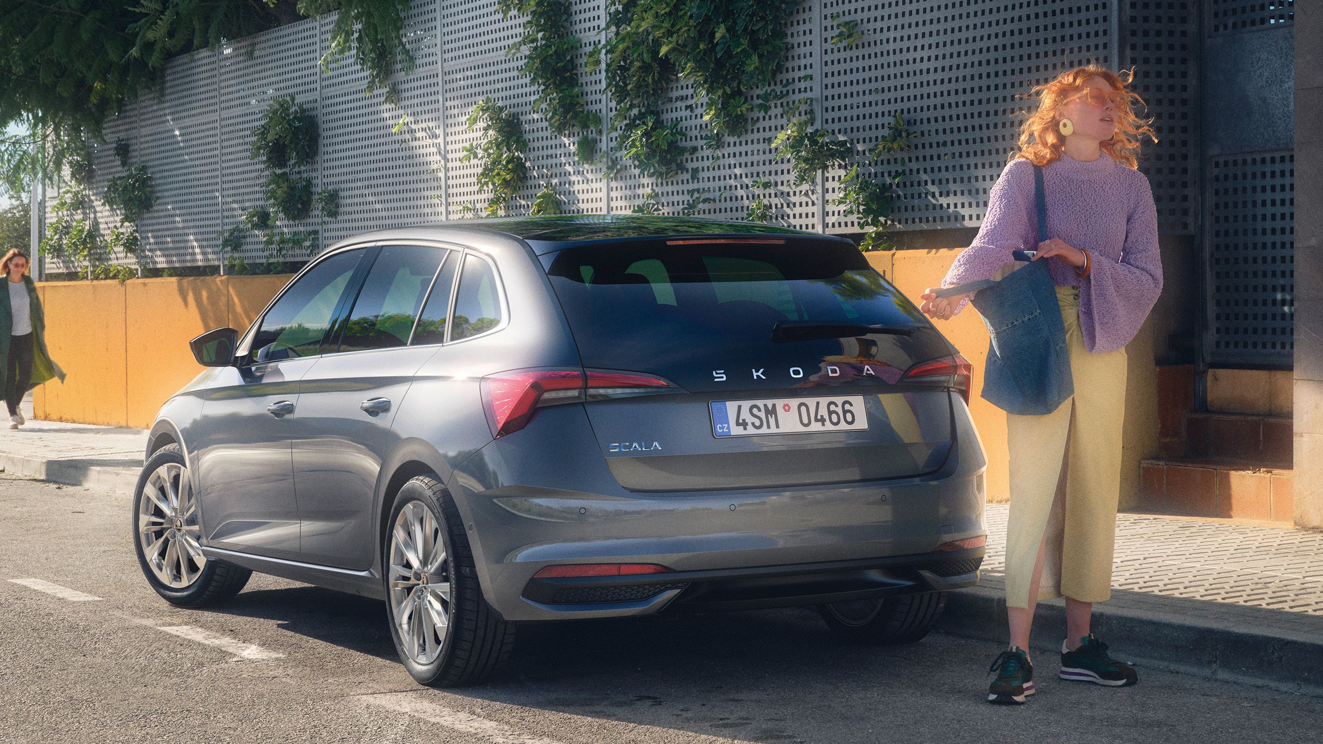 Oseba stoji ob parkiranem sivem avtomobilu znamke Škoda. Na steni za osebo je rastlinje. Oseba drži torbo in nosi svetlo vijolično bluzo ter rumene hlače.