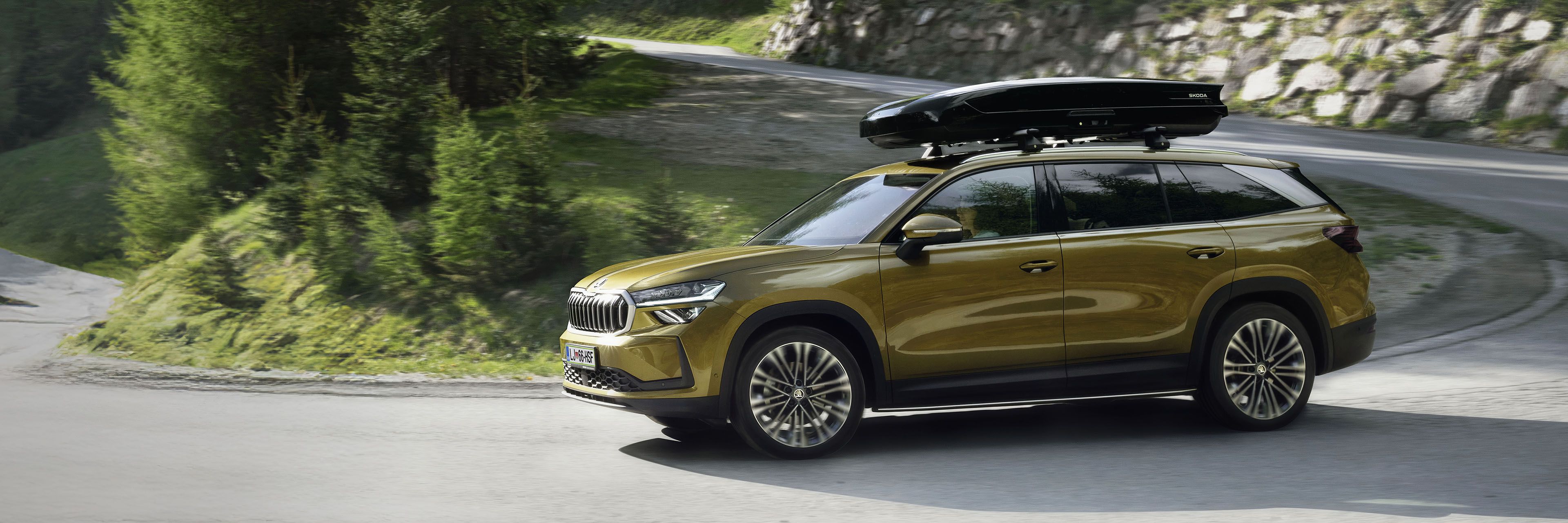 Avtomobil zlatkaste barve Škoda Kodiaq vozi po ovinkasti cesti obdan z drevesi in skalami. Na strehi avtomobila je pritrjena črna prtljažna škatla. Scena prikazuje naravo sredi dneva.