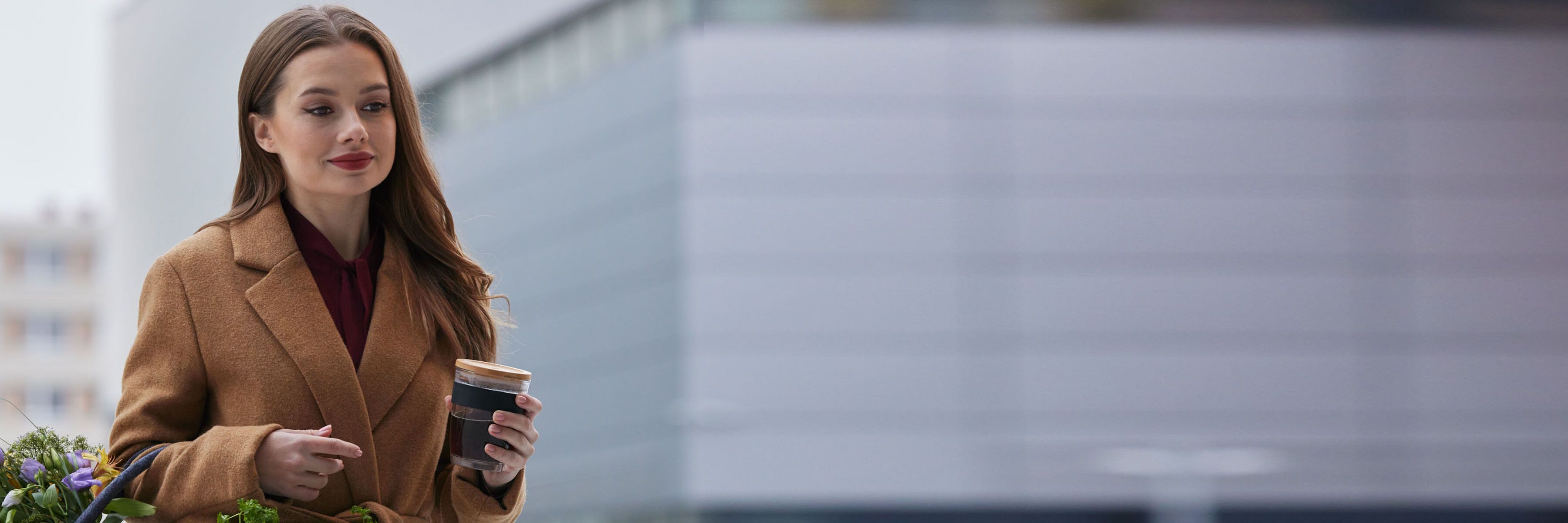 Oseba z rjavimi dolgimi lasmi, oblečena v rjav plašč, drži lonček za kavo in košaro. V ozadju je sodobna zgradba.