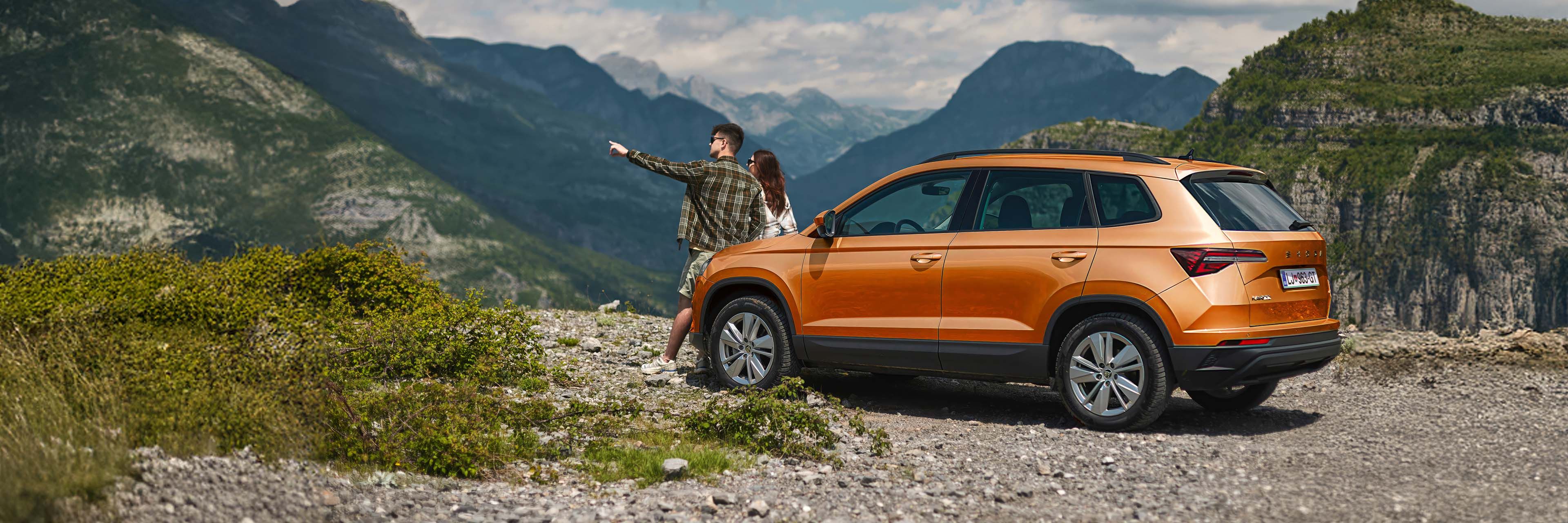 Svetlo oranžen SUV Škoda Karoq je parkiran ob robu ceste na gorskem območju. Dva človeka stojita ob vozilu; eden od njiju kaže proti oddaljenim goram. V ozadju se vidijo veličastni gorski vrhovi.