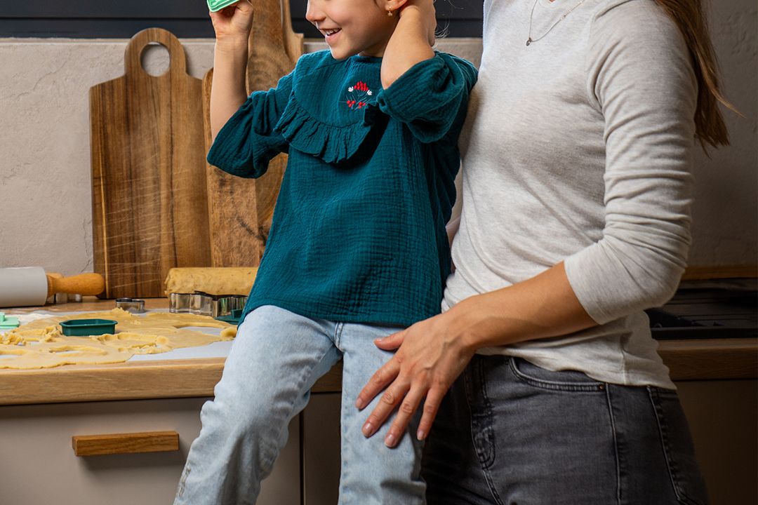 Ženska in deklica z zamegljenimi obrazi v kuhinji, kjer oblikujeta testo na pultu. Deklica drži zelen modelčki, medtem ko ženska nežno drži deklico za pas. V ozadju so lesene deske za rezanje in okno s pogledom na naravo.