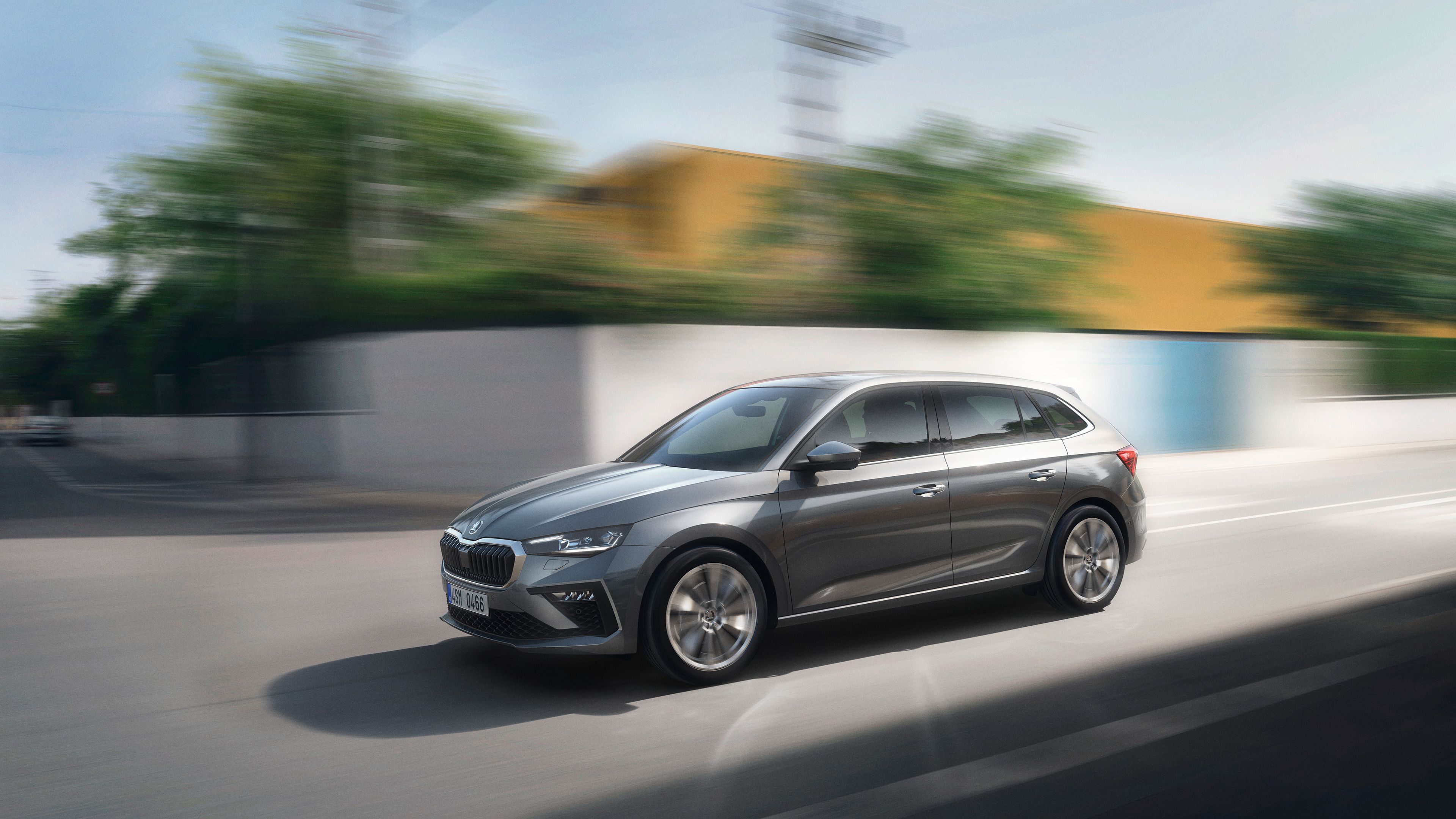 Siva Škoda Scala v gibanju na mestni cesti. Avtomobil je štirivratni, kompaktni model v kovinski barvi. Ozadje je zamegljeno zaradi hitrosti.